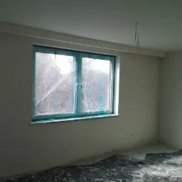 Pokój w trakcie remontu: ściany po szpachlowaniu, okno zabezpieczone folią, podłoga przykryta plandeką, wiszące kable.