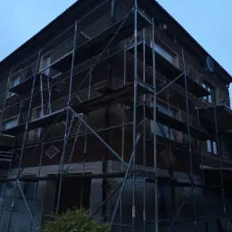 Dwupiętrowy budynek w trakcie prac elewacyjnych, osłonięte okna folią, rozstawione rusztowania, widoczne elementy dekoracyjne na elewacji w kształcie rombów.