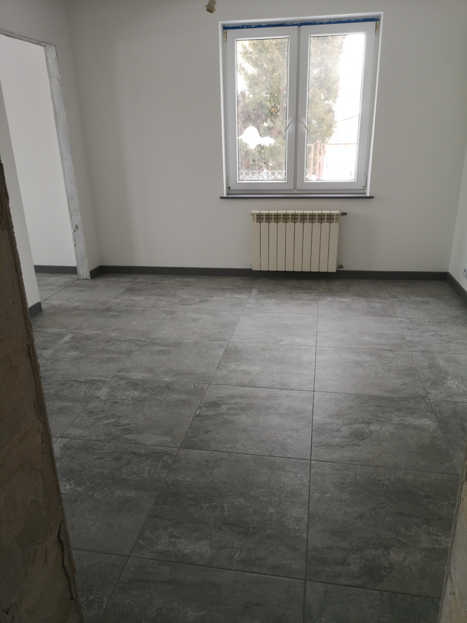 Wnętrze pokoju po remoncie z szarymi płytkami podłogowymi, białymi ścianami i grzejnikiem pod oknem. Widoczny fragment surowego muru przy wejściu.