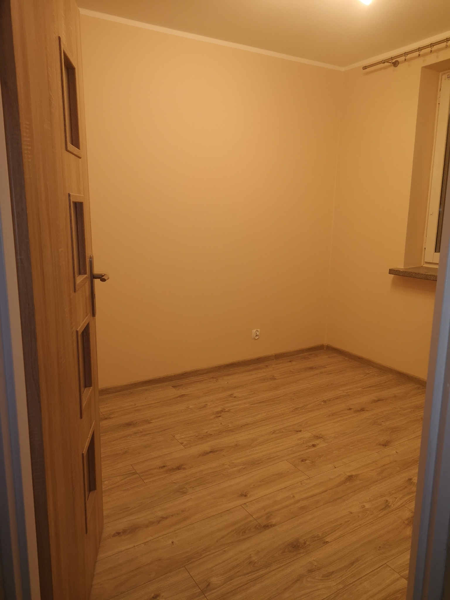 Pusty pokój z nową podłogą z paneli, świeżo pomalowanymi ścianami w odcieniach beżu i częściowo otwartymi drewnianymi drzwiami z przeszkleniami.