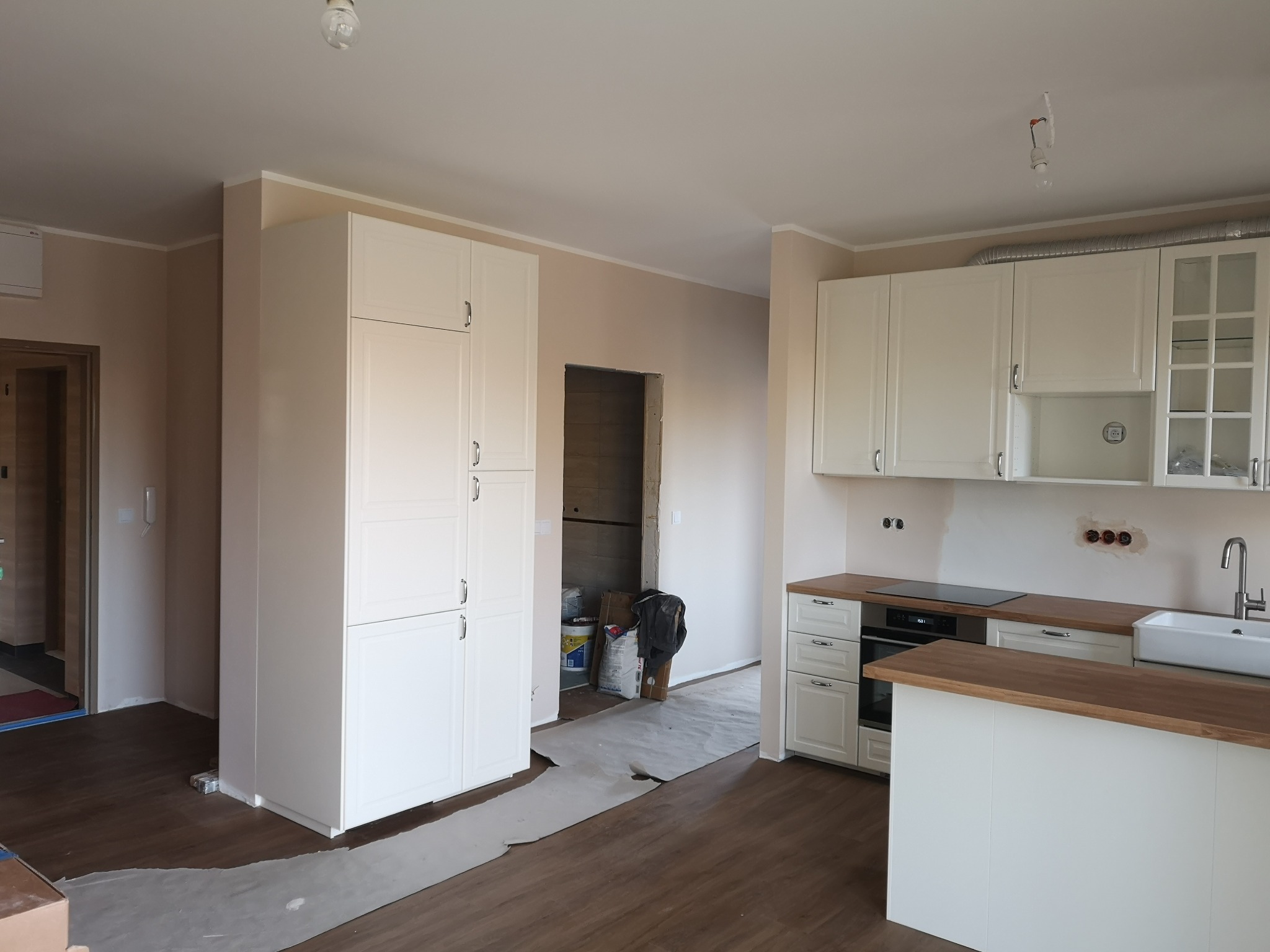 Wnętrze kuchni w trakcie montażu zabudowy meblowej, widoczne białe szafki z drewnianymi blatami, podłoga wyłożona panelami oraz ślady prac wykończeniowych na ścianach.
