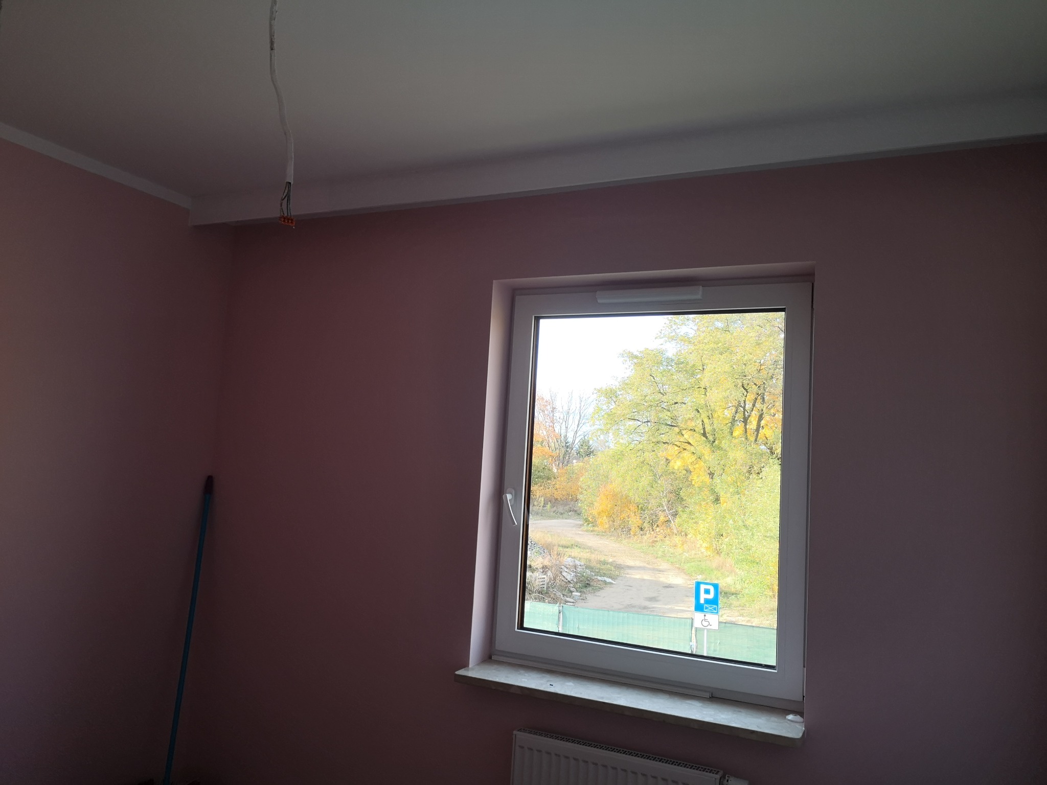 Pokój z różowymi ścianami w trakcie odświeżania, widoczne przewody elektryczne wystające z sufitu, okno z widokiem na jesienne drzewa i znak parkingowy.