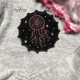 CNC Paweł Strzeszewski - Czarna naszywka z motywem łapacza snów, ozdobiona różowym haftem i błyszczącymi dżetami, umieszczona na szarym, dzierganym swetrze obok fragmentu różowej chusty z pomponami.