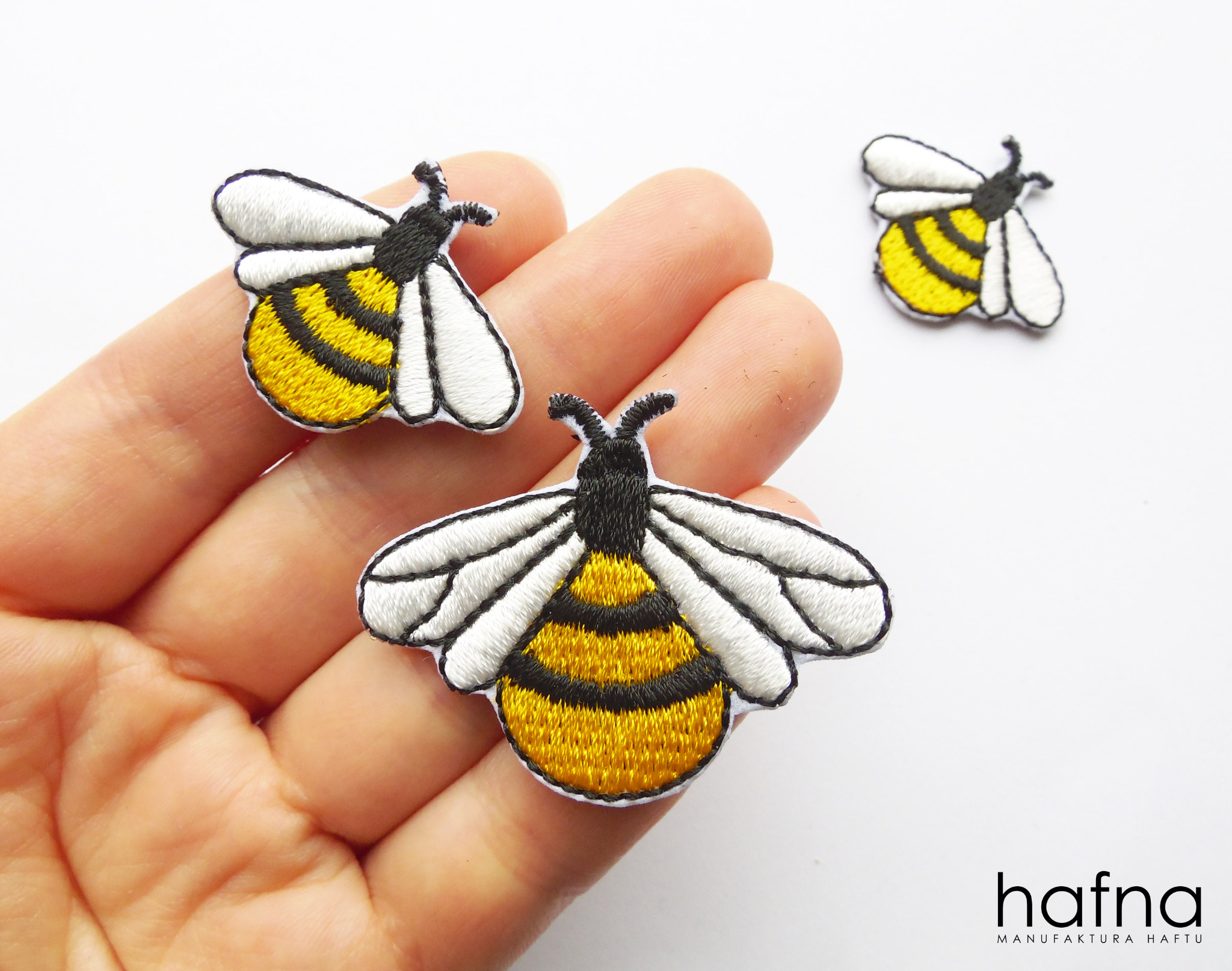 Trzy naszywki w kształcie pszczół na dłoni i białym tle, wykonane z precyzyjnego haftu, prezentujące detale żółto-czarnych pasków i białych skrzydeł.