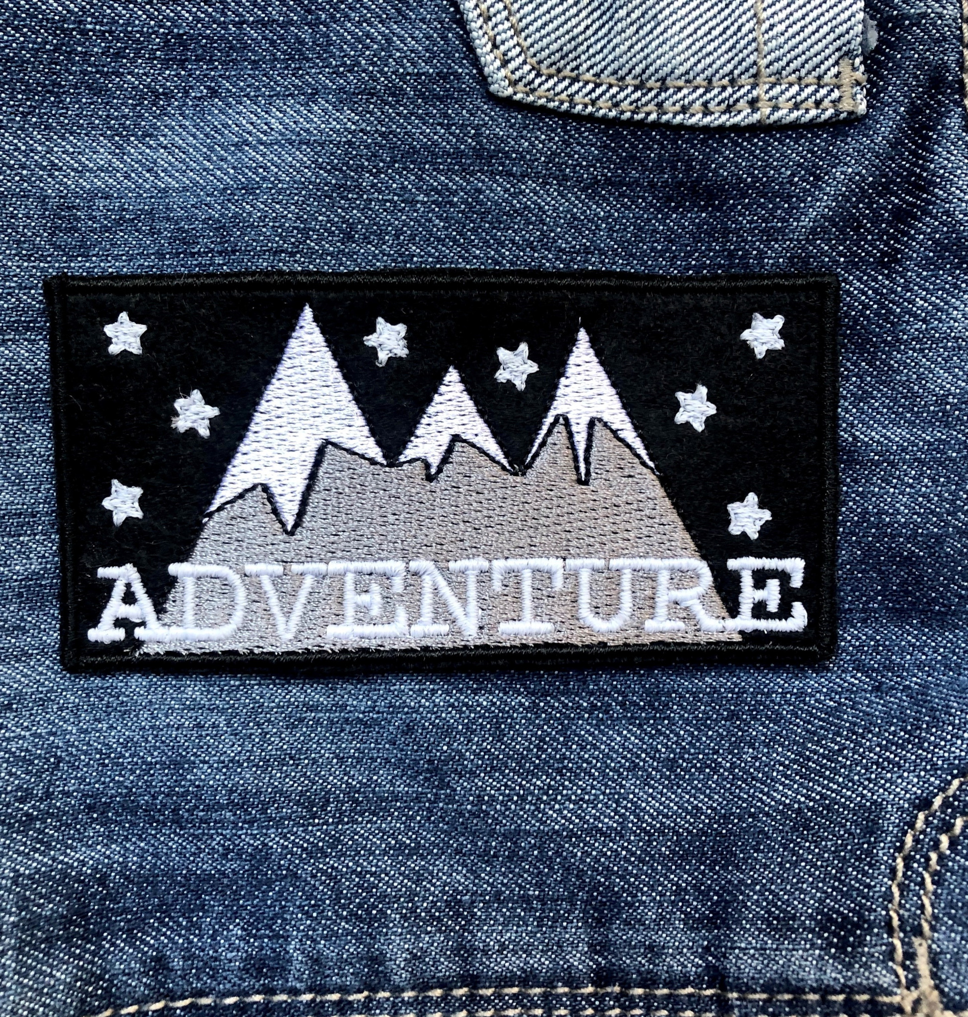 Naszywka z motywem gór, gwiazd i napisem 'Adventure' na tle dżinsowej kurtki, widoczne szwy i fragment kieszeni.