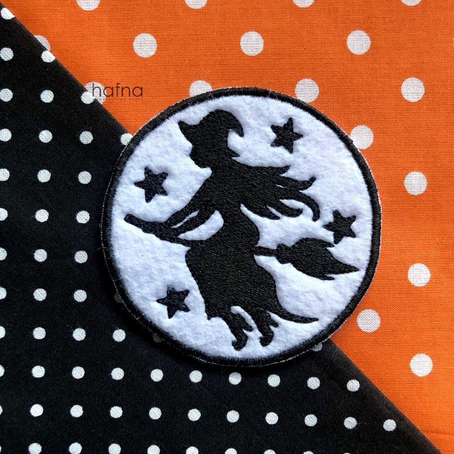 Okrągła naszywka z czarną sylwetką wiedźmy na miotle i białymi gwiazdami, na tle z czarno-pomarańczowej tkaniny w białe kropki.
