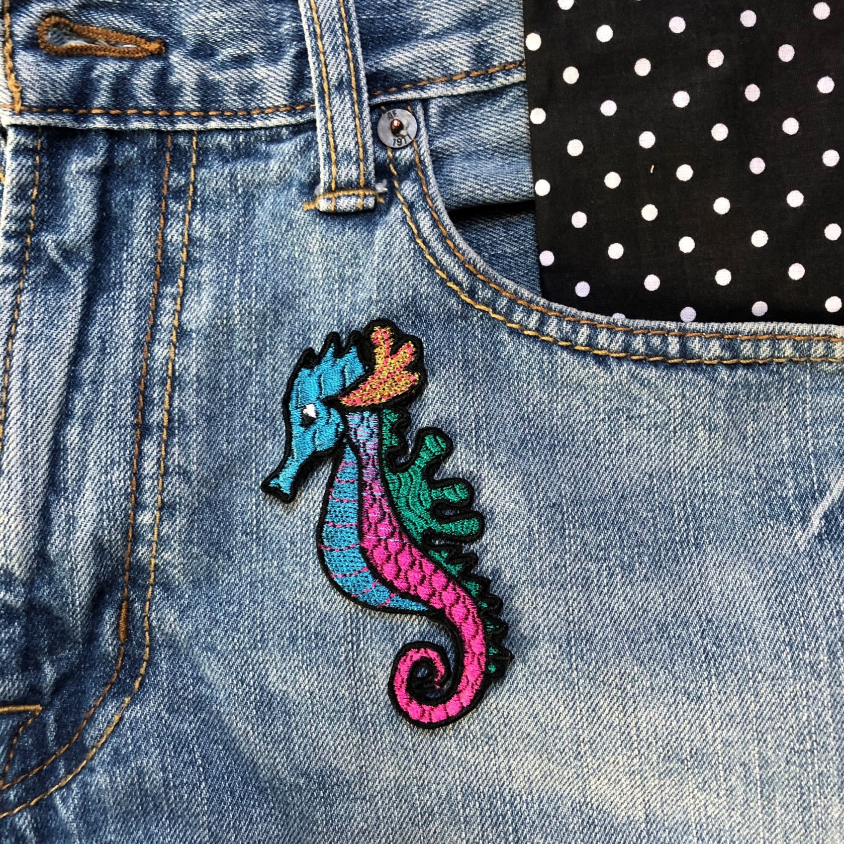 Naszywka z motywem kolorowego konika morskiego na kieszeni dżinsowej, z fragmentem materiału w kropki wystającym z kieszeni.