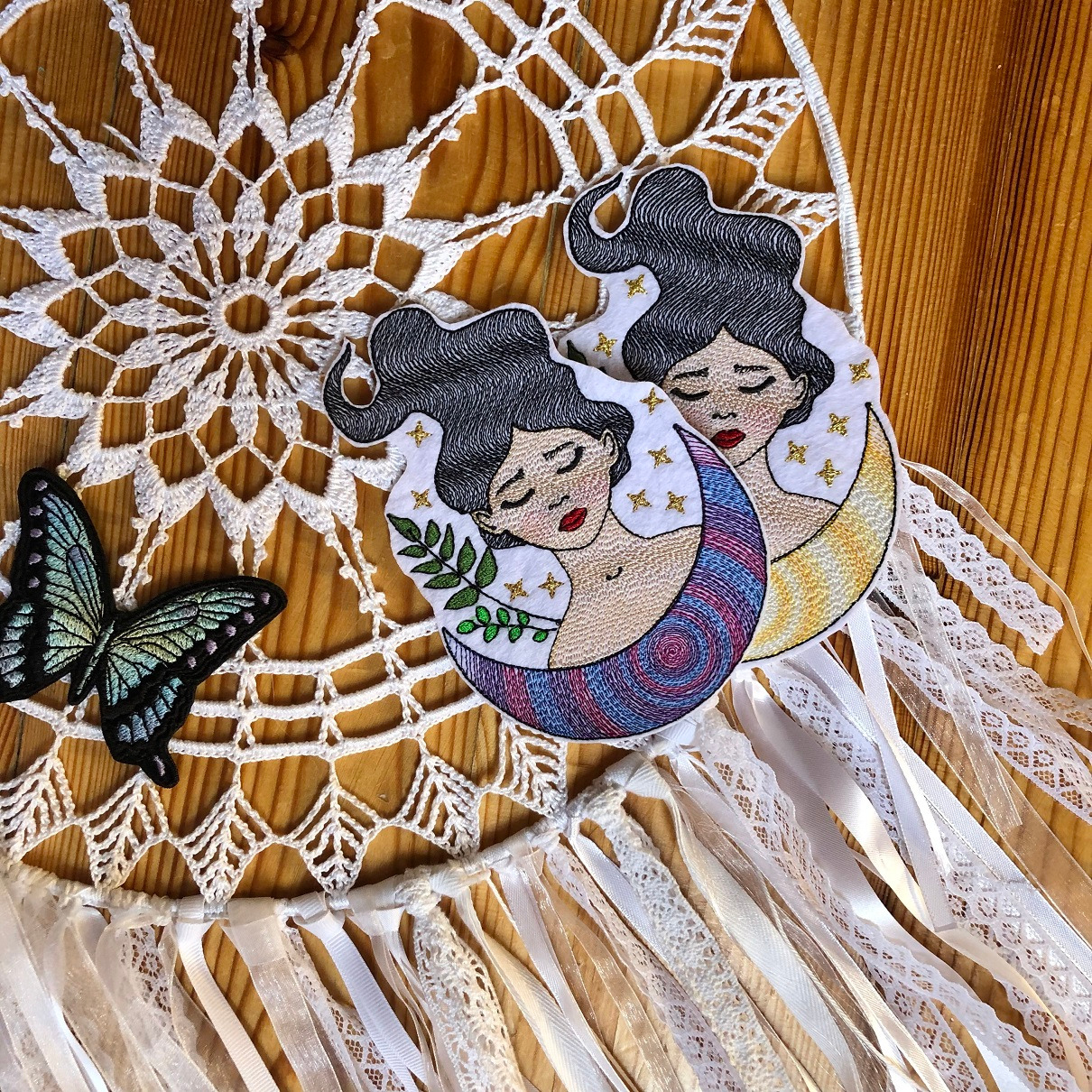 Ręcznie robiony łapacz snów z haftowanymi aplikacjami przedstawiającymi kobiety na księżycach, z dodatkiem koronki, wstążek i haftowanego motyla na drewnianym tle.
