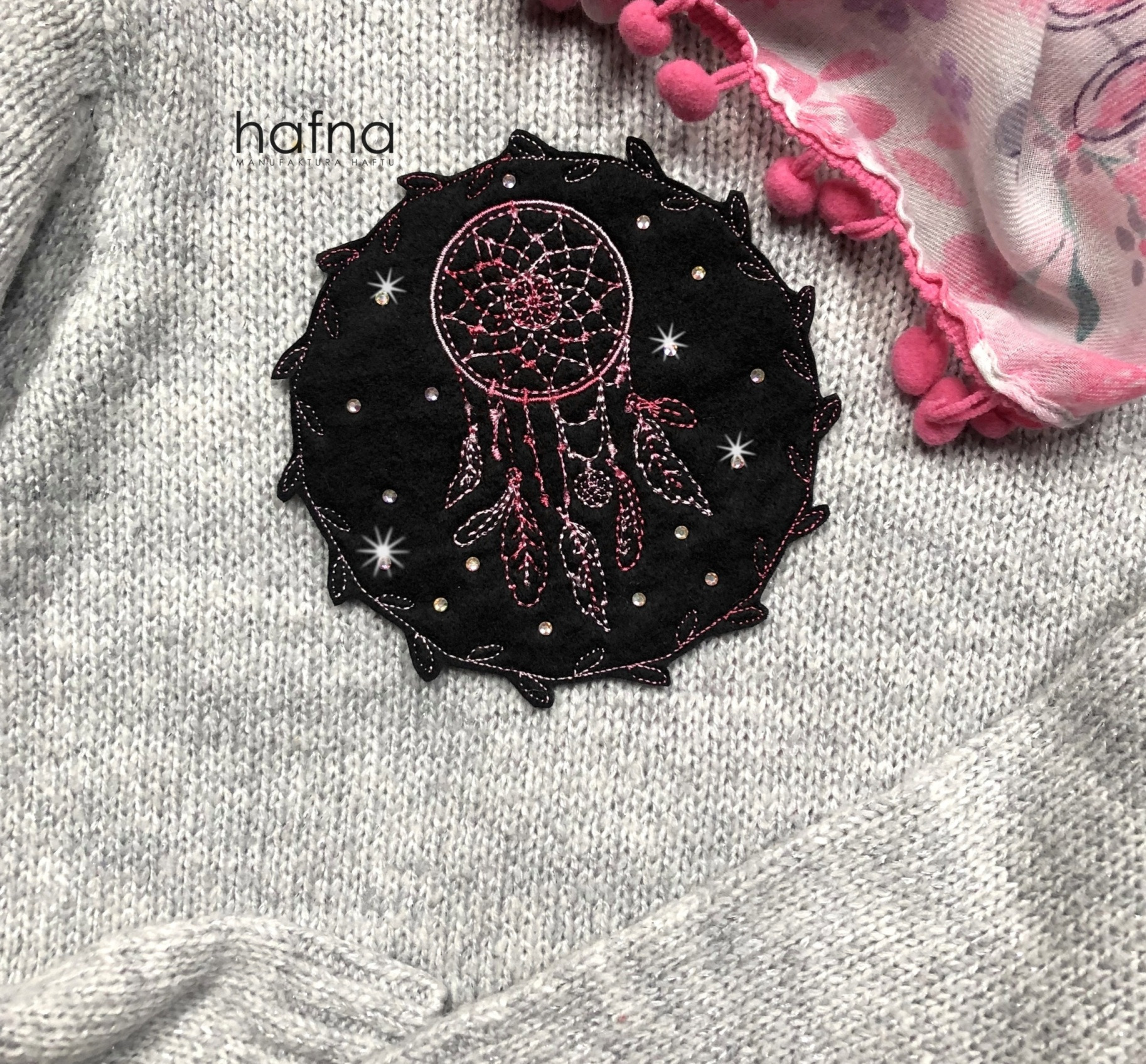 Czarna naszywka z motywem łapacza snów, ozdobiona różowym haftem i błyszczącymi dżetami, umieszczona na szarym, dzierganym swetrze obok fragmentu różowej chusty z pomponami.
