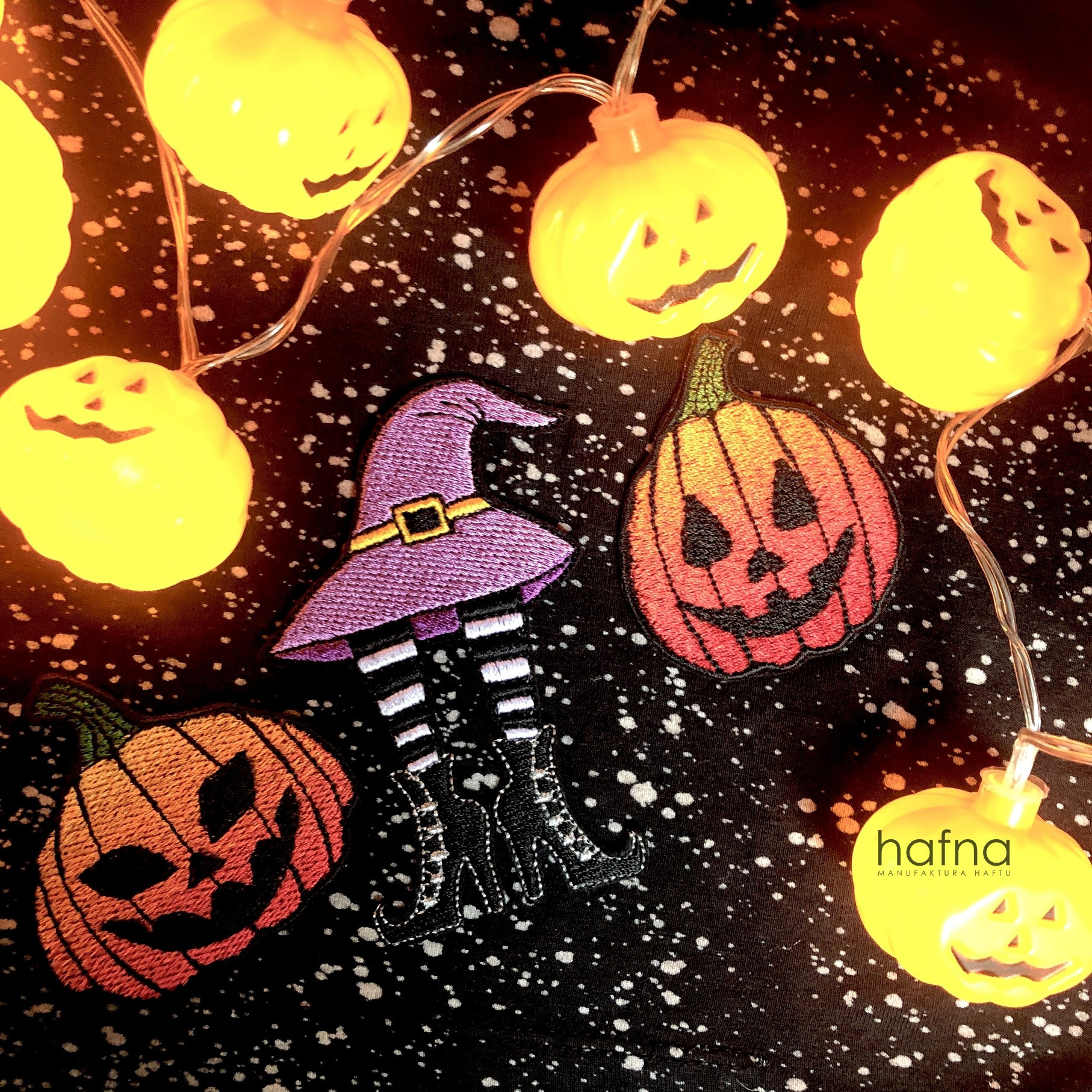 Halloween themed embroidered patches of pumpkins and a witch's hat with striped legs, surrounded by pumpkin-shaped string lights on a speckled black fabric, showcasing detailed stitching.
