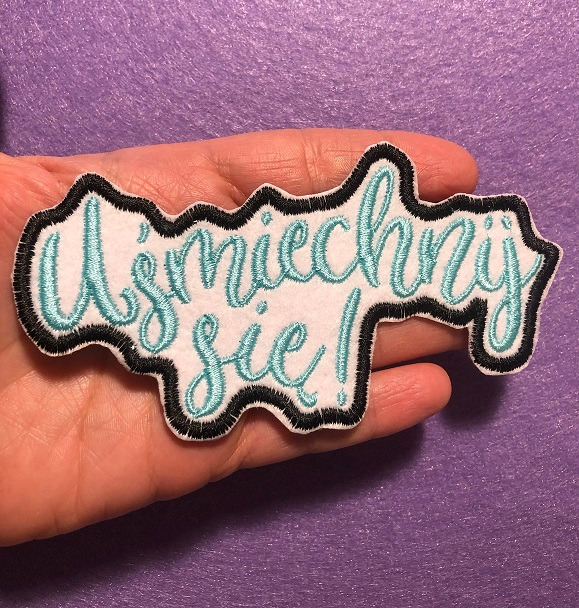 Naszywka z wyhaftowanym napisem 'Uśmiechnij się!' w kolorze turkusowym, z czarną obwódką, trzymana w dłoni na fioletowym tle.