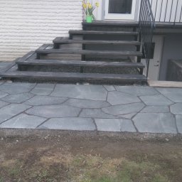 KAMSTEN kamieniarstwo budowlane - Nieregularny kształt szarej kamiennej nawierzchni prowadzącej do drewnianych schodów z metalową barierką i wejścia do piwnicy.