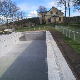 KAMSTEN kamieniarstwo budowlane - Niedokończony basen wyłożony jasnoszarą kamienną mozaiką, widok na żółty dom w tle i błękitne niebo z chmurami.