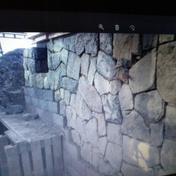 KAMSTEN kamieniarstwo budowlane - Wznoszenie ściany z nieregularnych kamieni, widoczne rusztowanie i przygotowane podłoże, w tle duży głaz.