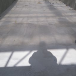 KAMSTEN kamieniarstwo budowlane - Wnętrze betonowego basenu w trakcie budowy, widoczne ściany i dno z szarym betonem, cień osoby fotografującej, liście na dnie, pomarańczowe pasy zabezpieczające na górze.