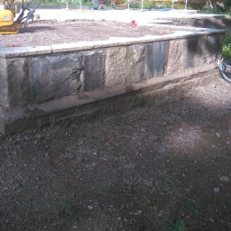 KAMSTEN kamieniarstwo budowlane - Budowa kamiennego murku oporowego z szarych kamieni, widoczny ubijak wibracyjny, konstrukcja zadaszenia i pracownik w tle.