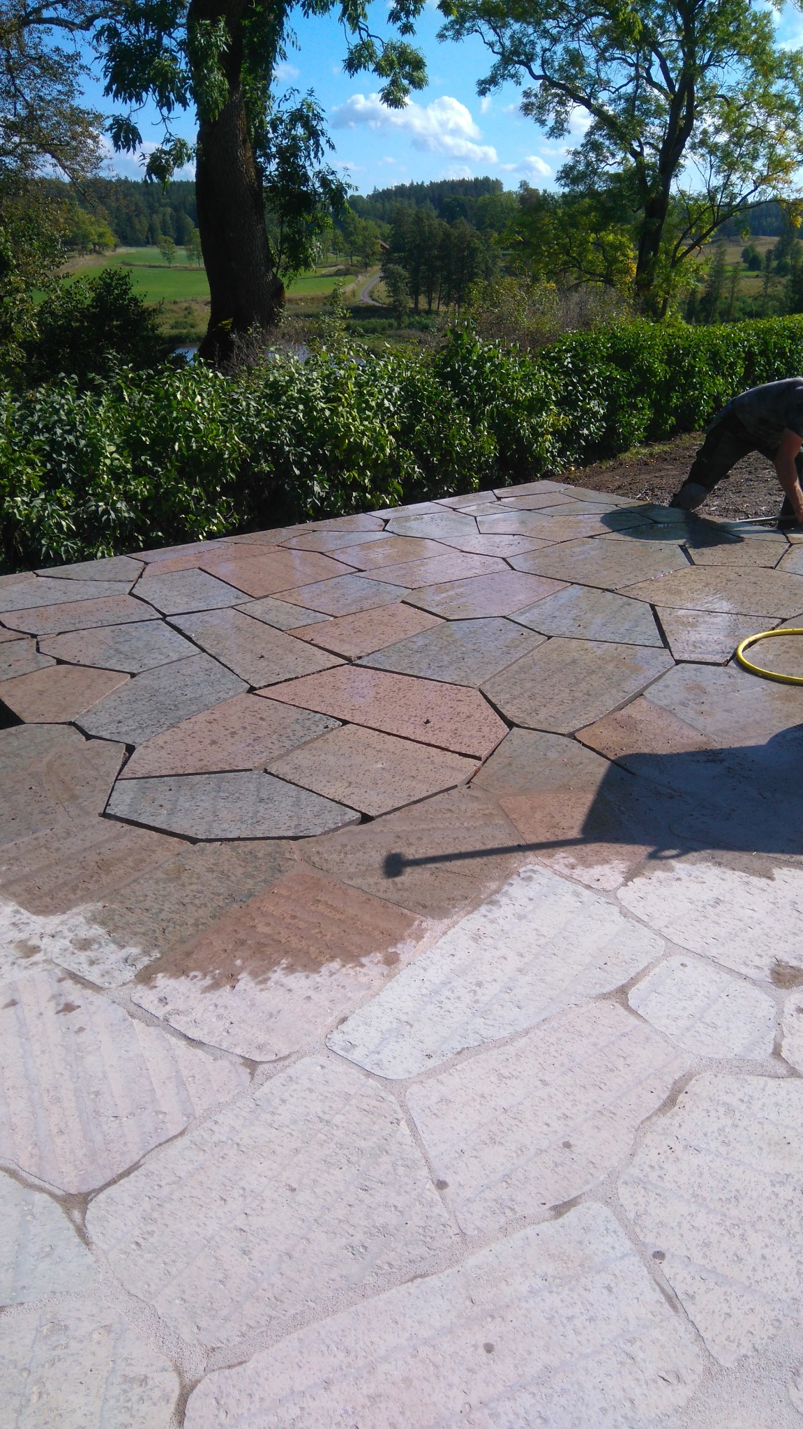 Układanie tarasu z nieregularnych płyt kamiennych w ogrodzie, widoczne fragmenty żółtego węża ogrodowego i sylwetka pracownika, zieleń w tle.