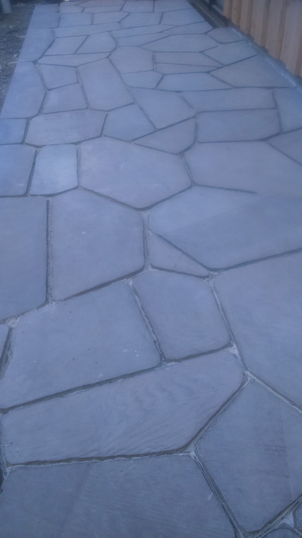 Nieregularne, szare płyty kamienne tworzące ścieżkę lub taras, z fugami wypełnionymi jasnym materiałem, widziane pod kątem.