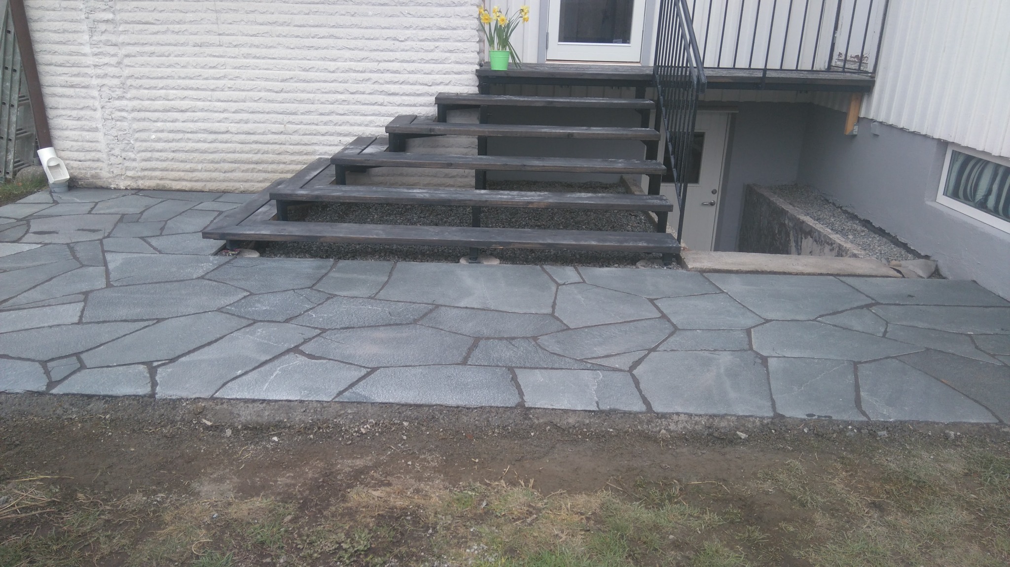 Nieregularny kształt szarej kamiennej nawierzchni prowadzącej do drewnianych schodów z metalową barierką i wejścia do piwnicy.