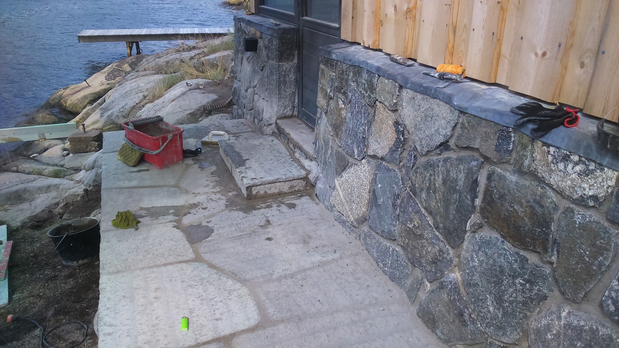 Budowa kamiennego tarasu przy budynku z drewnianą elewacją, widoczne narzędzia i materiały budowlane, w tle skały i woda.