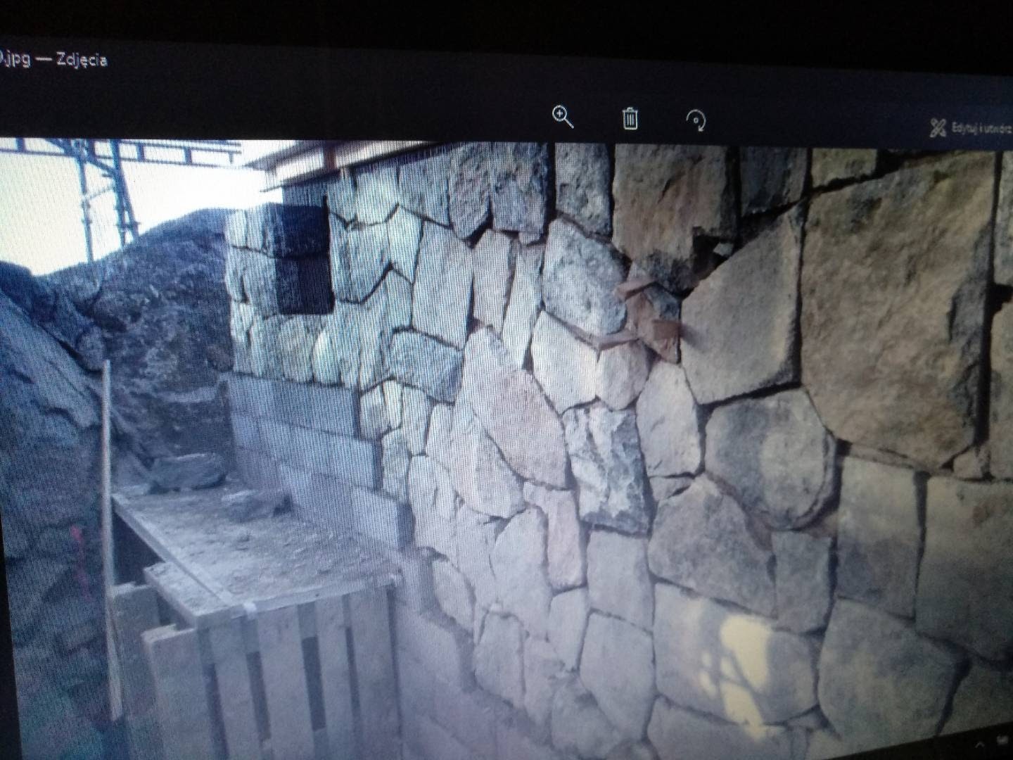 Wznoszenie ściany z nieregularnych kamieni, widoczne rusztowanie i przygotowane podłoże, w tle duży głaz.