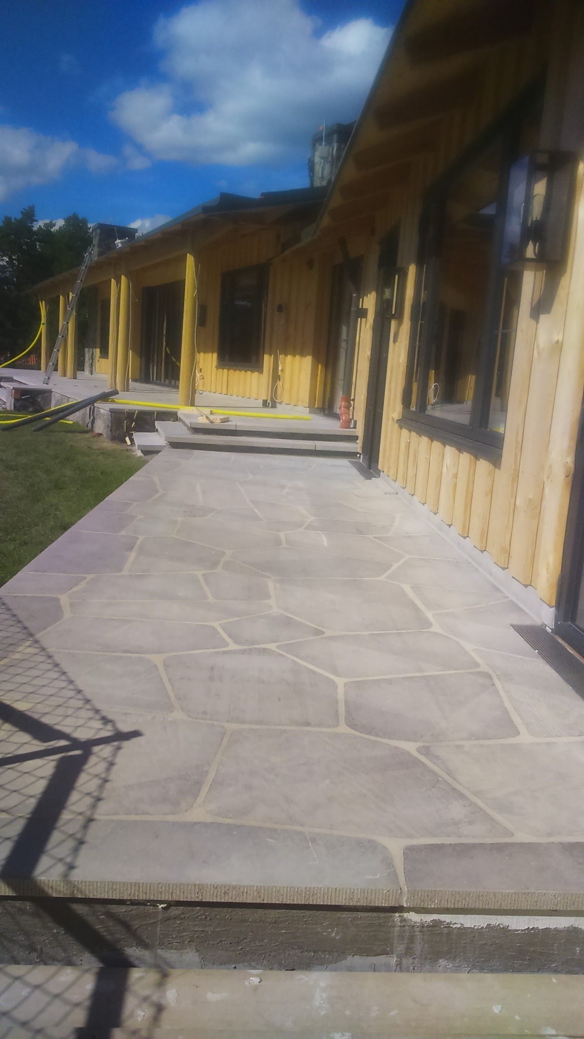 Jasnoszara kamienna ścieżka z nieregularnych płyt prowadząca wzdłuż drewnianego budynku z kolumnami, widoczne schody i trawnik.