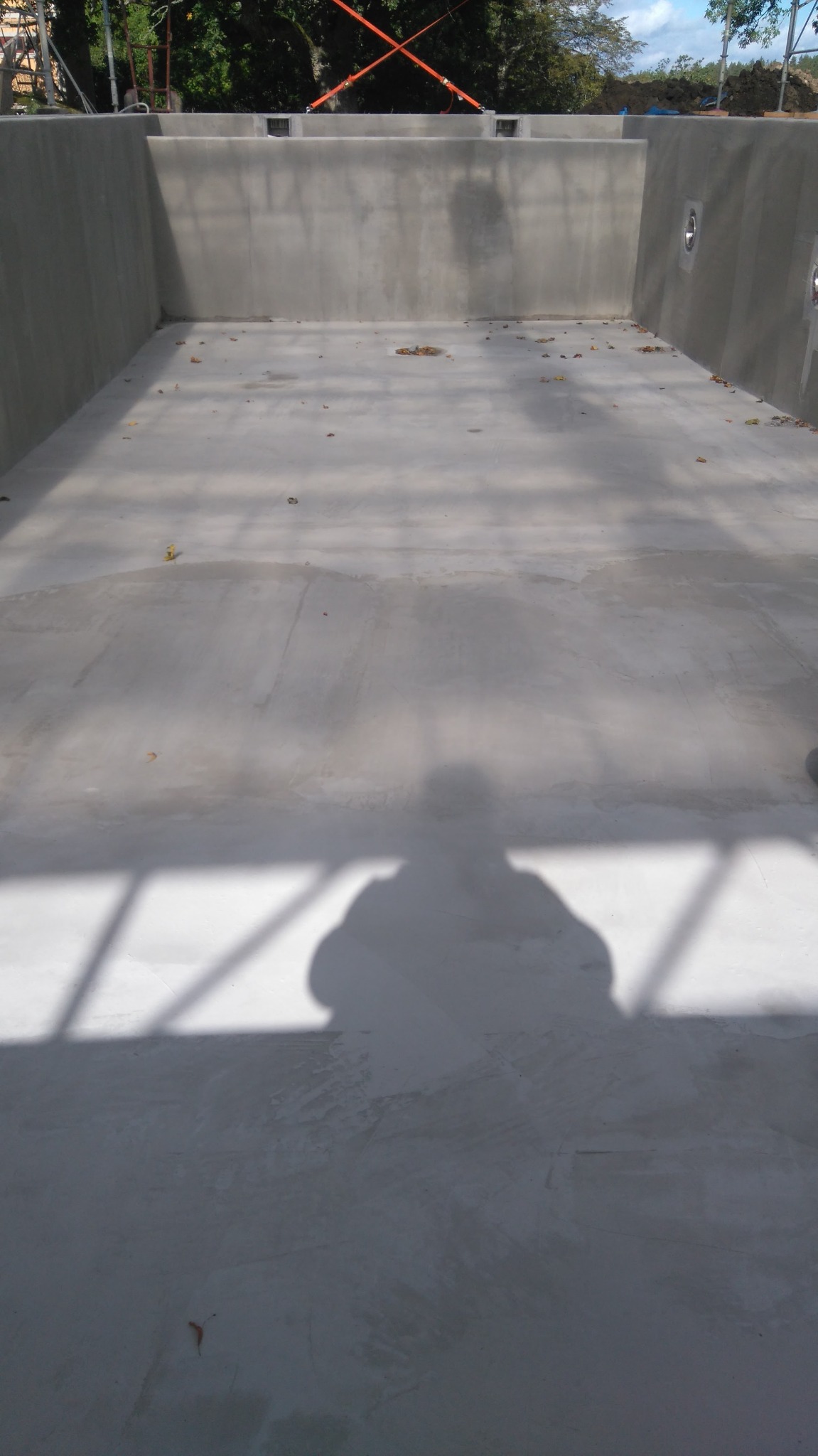 Wnętrze betonowego basenu w trakcie budowy, widoczne ściany i dno z szarym betonem, cień osoby fotografującej, liście na dnie, pomarańczowe pasy zabezpieczające na górze.