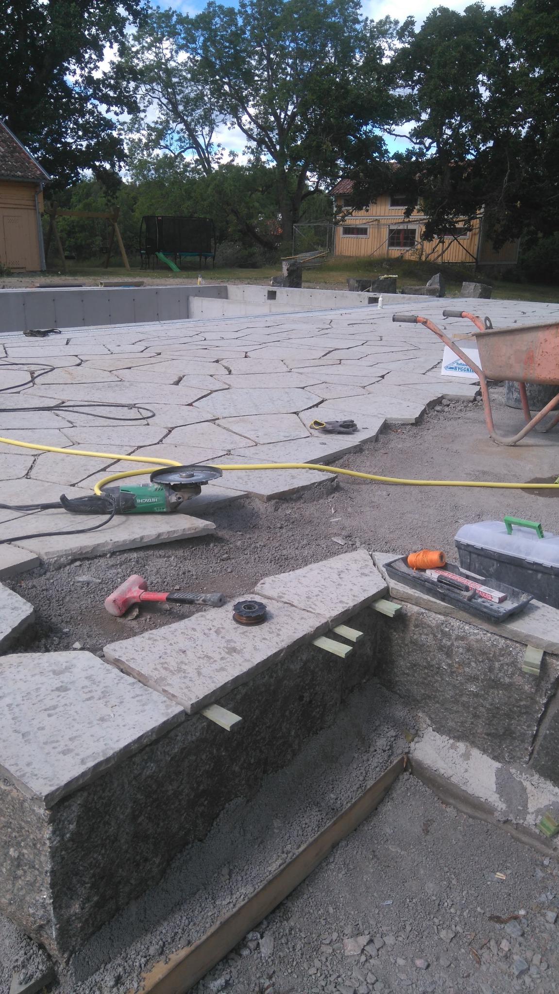 Układanie kamiennego tarasu z użyciem szlifierki kątowej i poziomicy, widoczny wózek transportowy w tle.