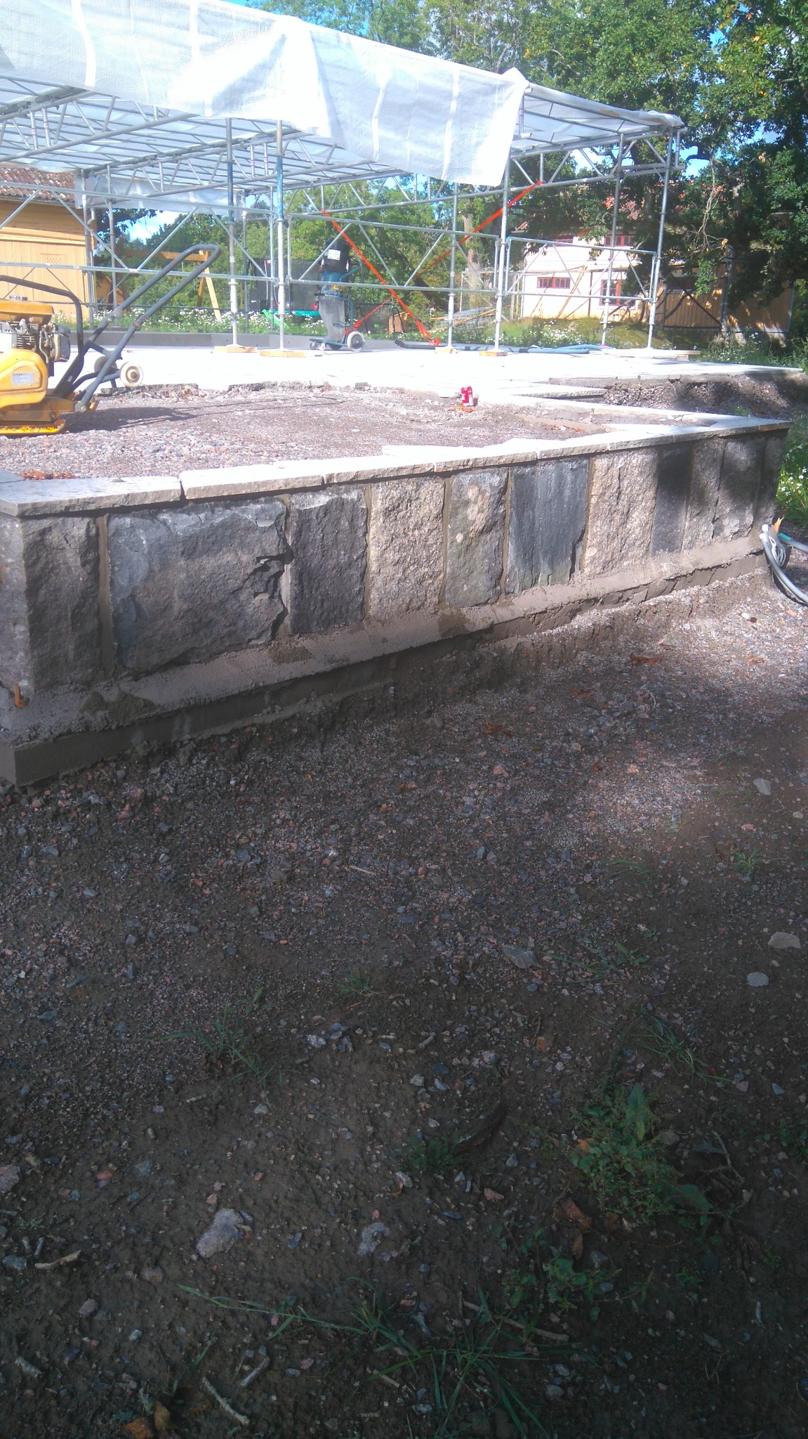 Budowa kamiennego murku oporowego z szarych kamieni, widoczny ubijak wibracyjny, konstrukcja zadaszenia i pracownik w tle.