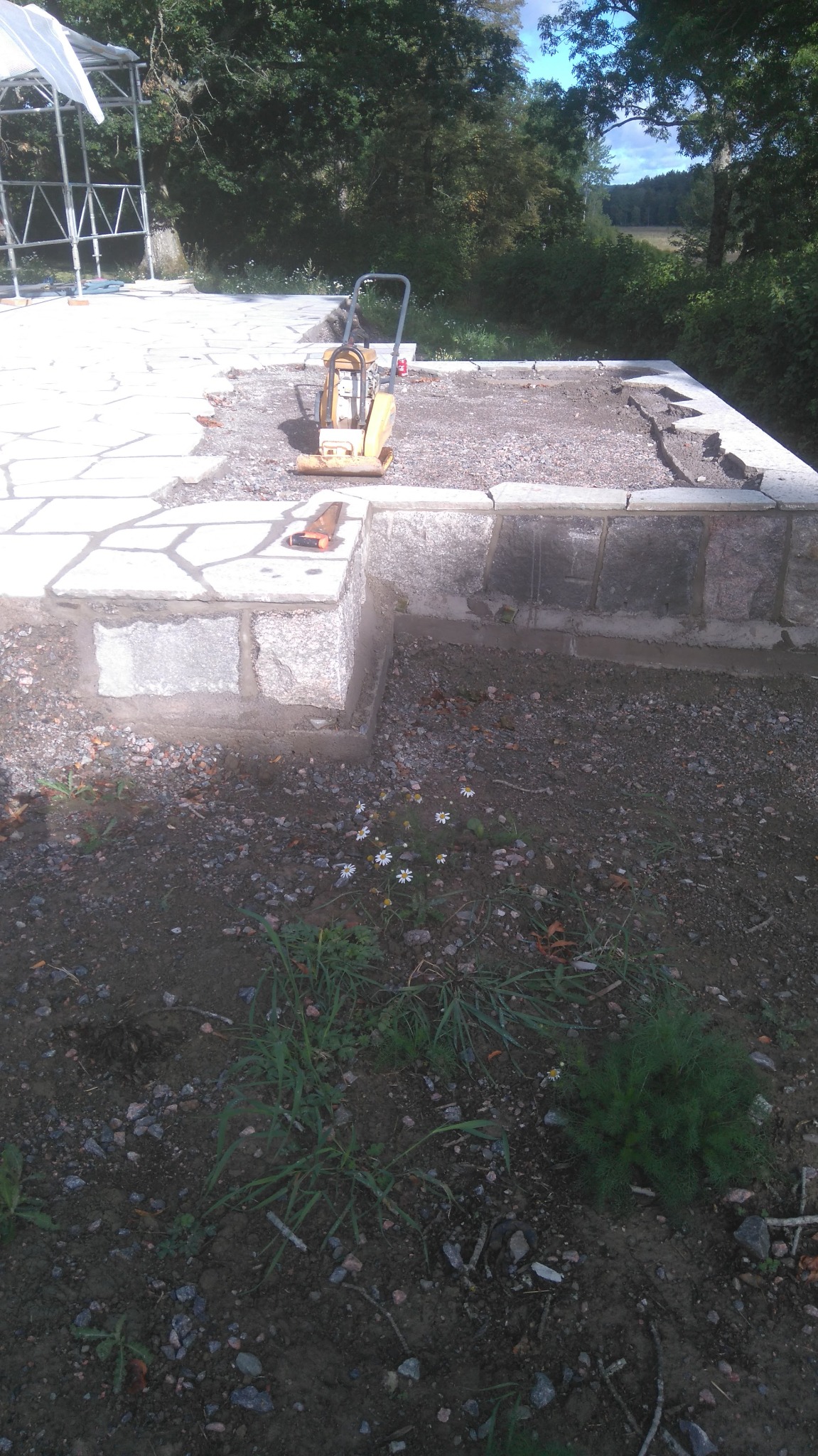 Żółta zagęszczarka wibrująca na podsypce pod taras z nieregularnych płyt kamiennych, widoczne fundamenty i zarys konstrukcji zadaszenia.