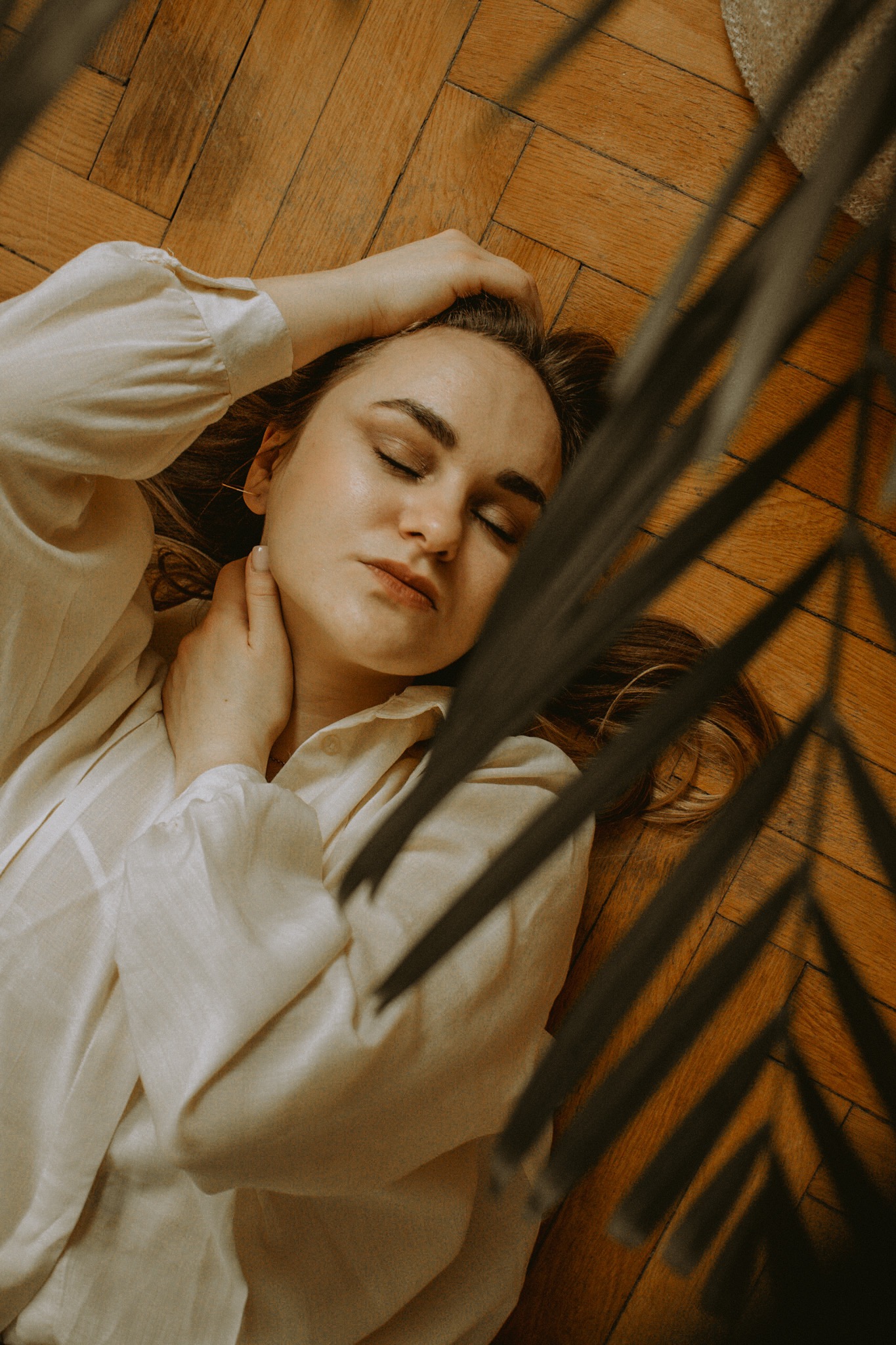 Portret kobiety leżącej na drewnianej podłodze w jasnej koszuli, z zamkniętymi oczami i dłonią na szyi, z dekoracyjnymi liśćmi w kadrze.