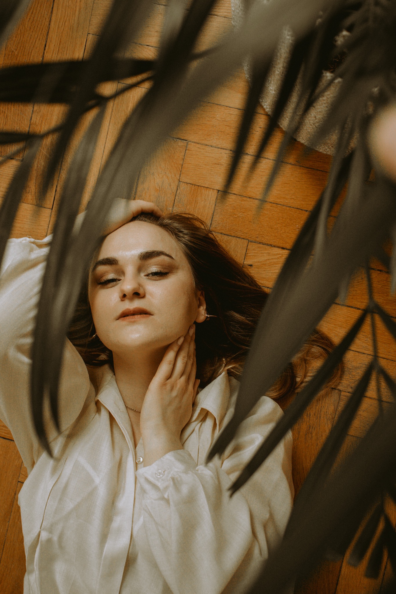 Portret kobiety leżącej na drewnianej podłodze, ujęcie z góry przez liście rośliny doniczkowej, złote kolczyki i delikatny makijaż.