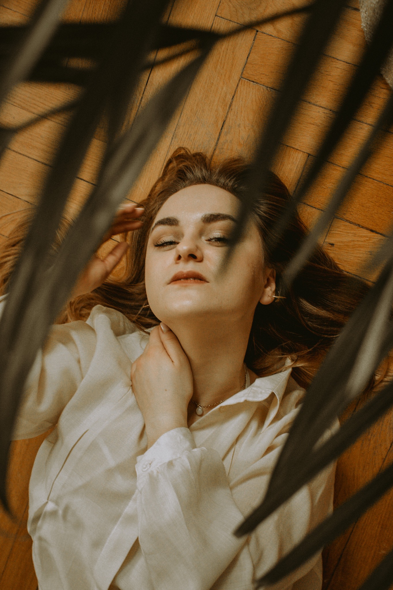 Kobieta leżąca na drewnianej podłodze w jasnej koszuli, ujęcie z góry przez liście rośliny, delikatny makijaż i subtelna biżuteria.