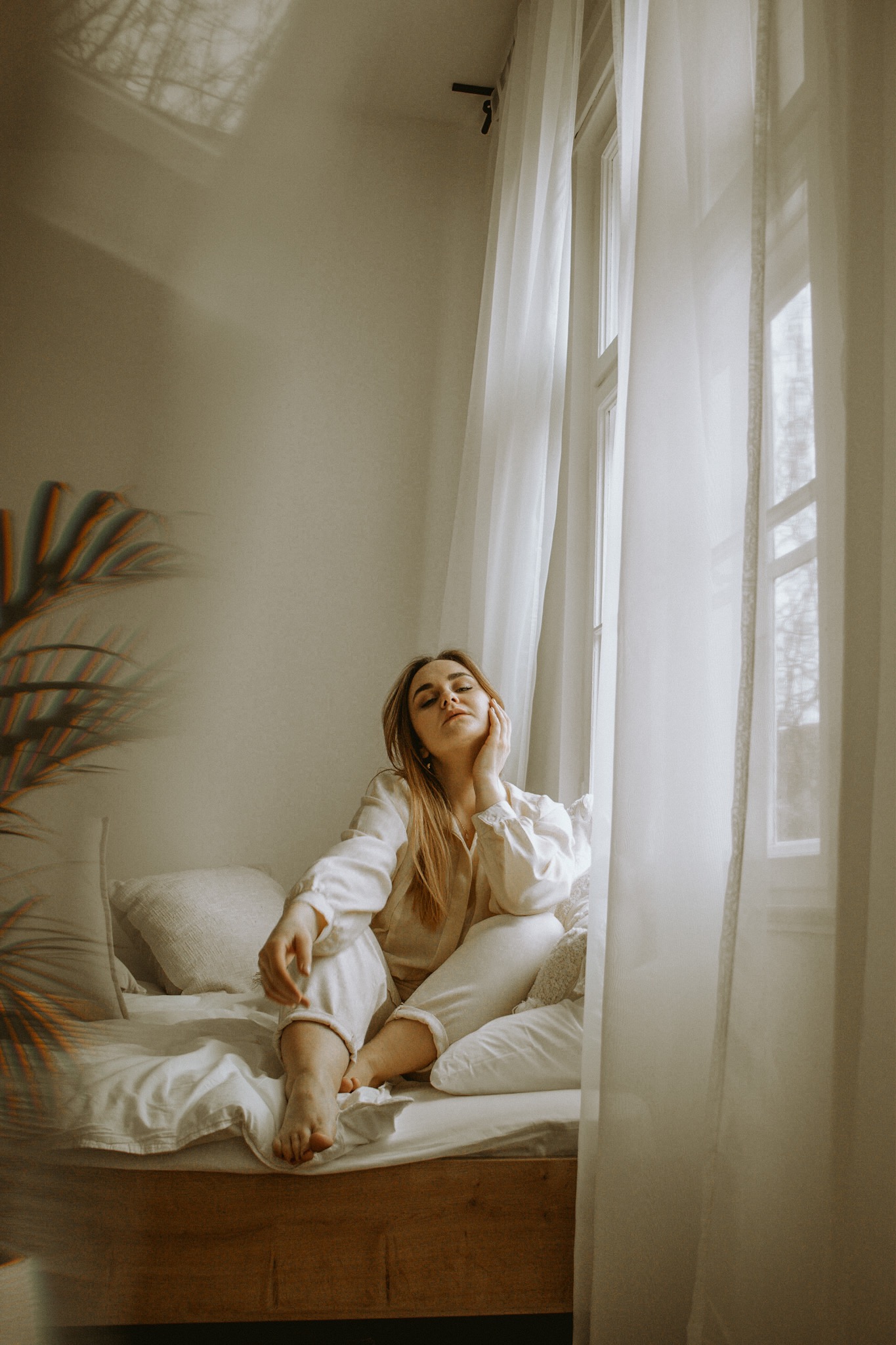Kobieta w jasnej piżamie siedzi na łóżku przy oknie z białymi zasłonami, zamyślona, w tle roślina doniczkowa.
