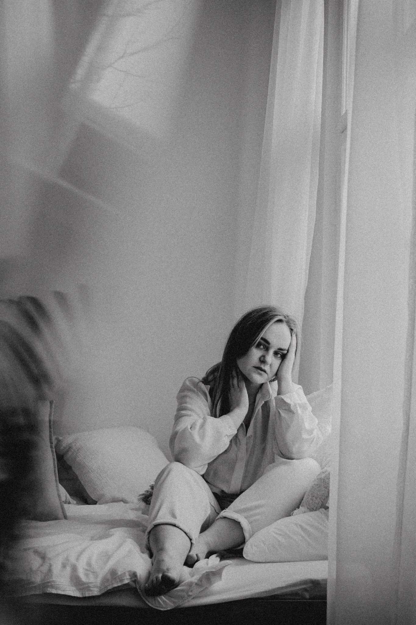 Kobieta w jasnym ubraniu siedzi na łóżku, opierając głowę na dłoniach, zamyślony wyraz twarzy, obok okno z delikatną zasłoną, monochromatyczne ujęcie.
