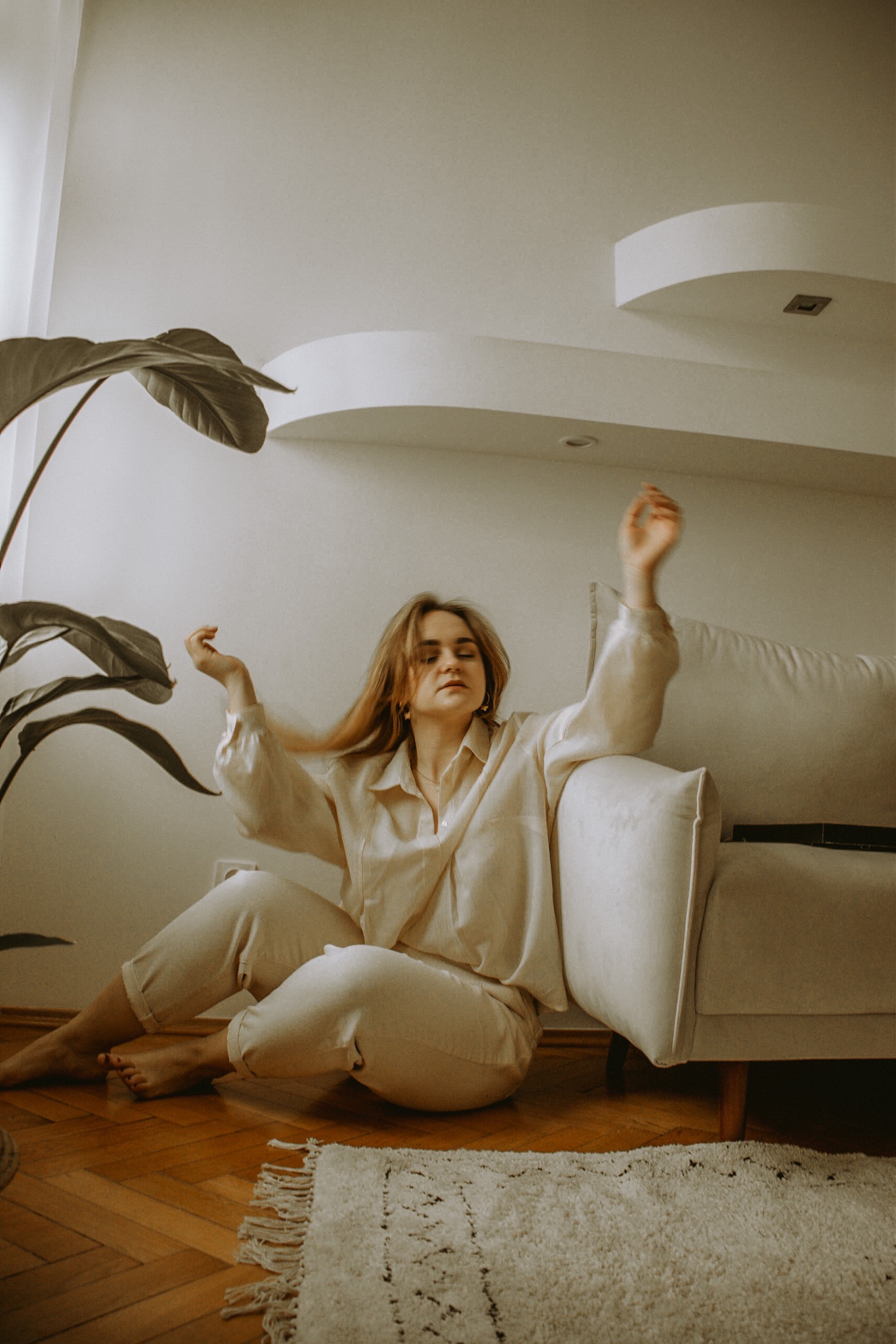Kobieta w beżowym komplecie siedzi na podłodze obok sofy, z rośliną doniczkową w tle, w minimalistycznym wnętrzu z drewnianą podłogą i białym dywanem.
