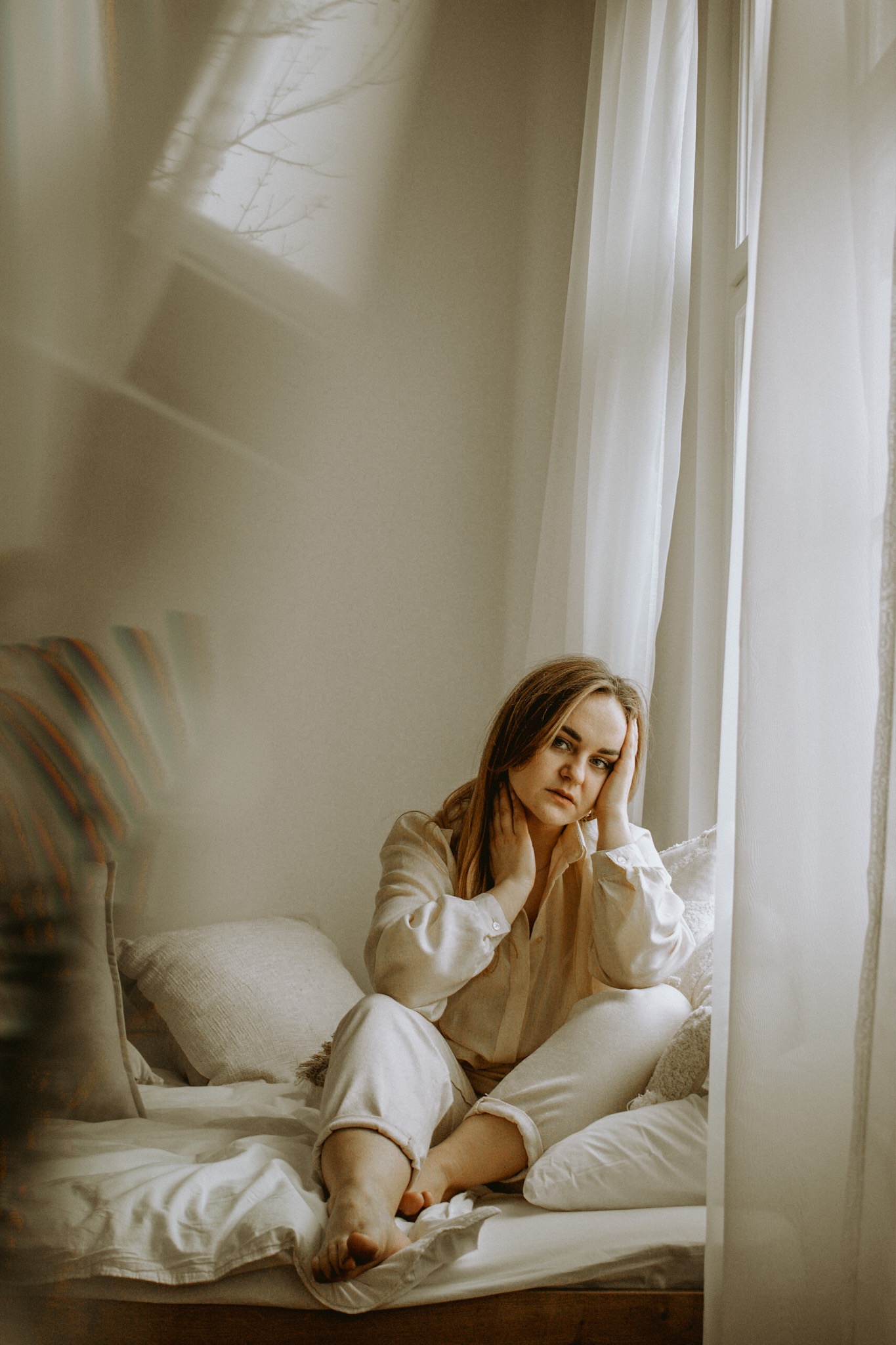 Portret kobiety w jasnym lnianym stroju siedzącej na łóżku przy oknie z delikatnymi zasłonami, zamyślony wyraz twarzy, naturalne światło.