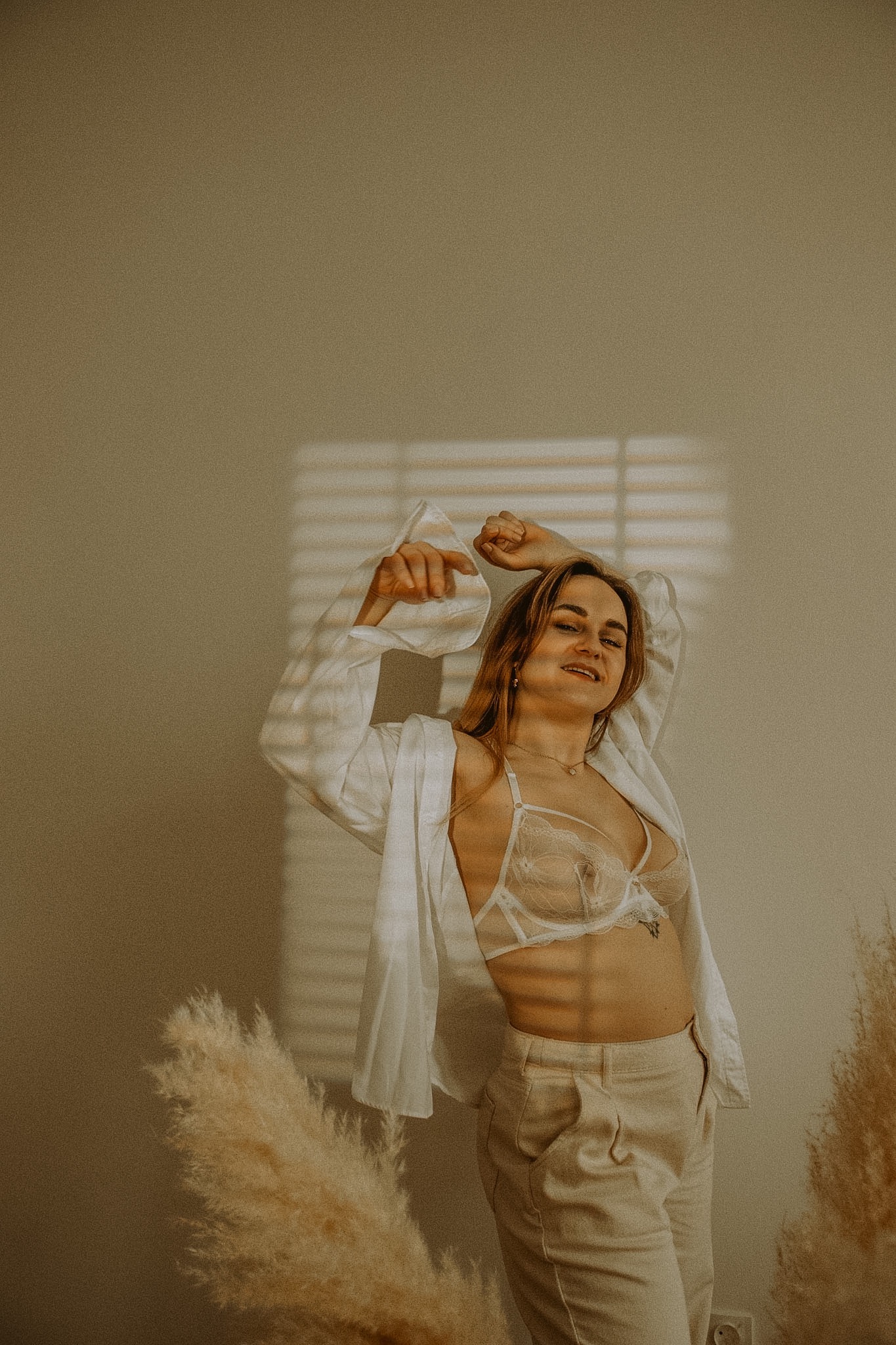 Portret kobiety w bieliźnie i białej koszuli, opierającej się o ścianę z cieniem żaluzji, obok dekoracyjnych traw pampasowych.
