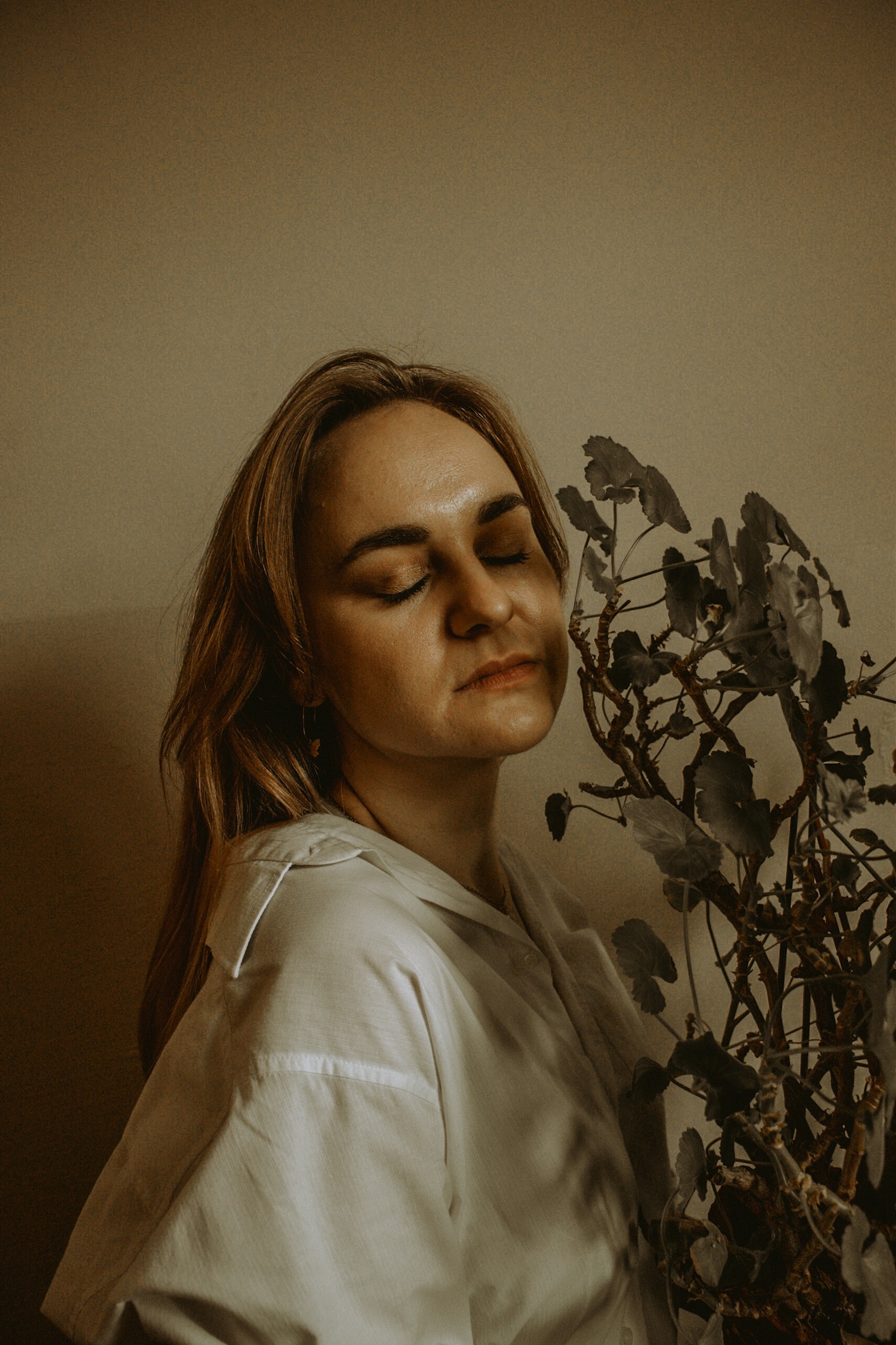 Portret kobiety z zamkniętymi oczami, w białej koszuli, stojącej obok rośliny doniczkowej na tle beżowej ściany, w ciepłym oświetleniu.