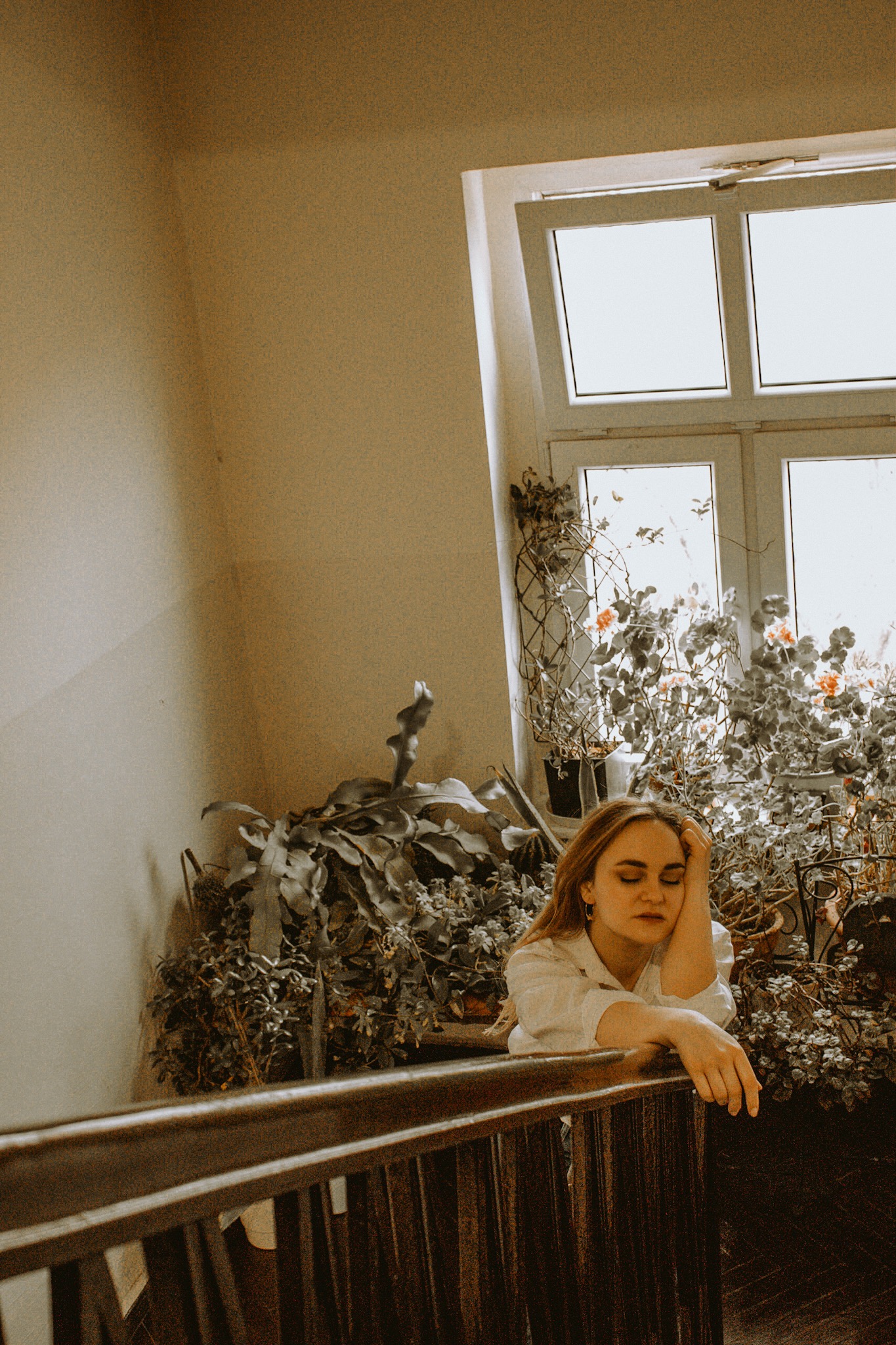 Kobieta z zamkniętymi oczami opiera się o drewnianą balustradę, za nią okno z dużą ilością roślin doniczkowych w stonowanych barwach.