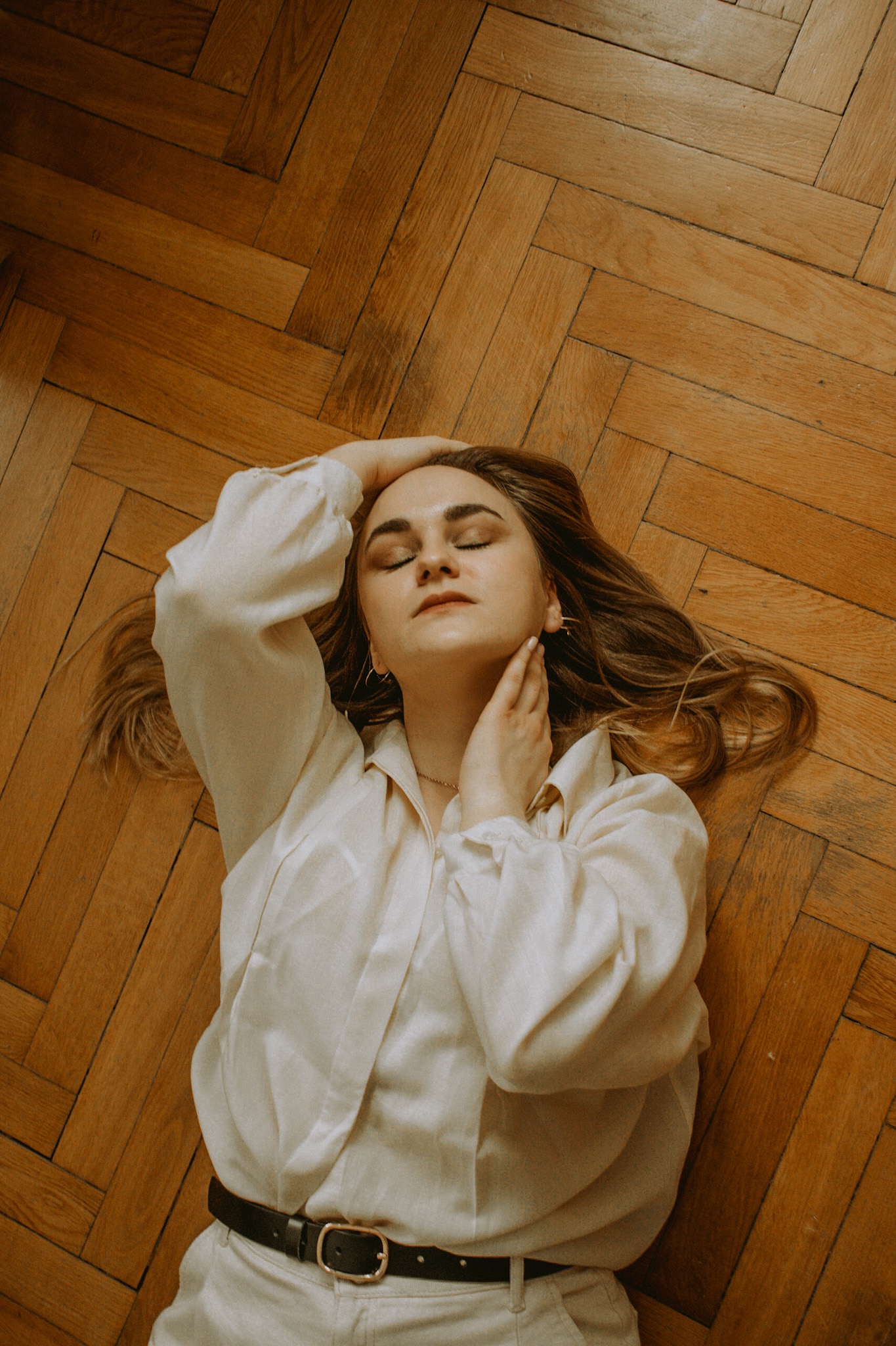 Kobieta w jasnej koszuli i spodniach, leżąca na parkiecie w jodełkę, z zamkniętymi oczami i dłonią na szyi, w spokojnej pozie.