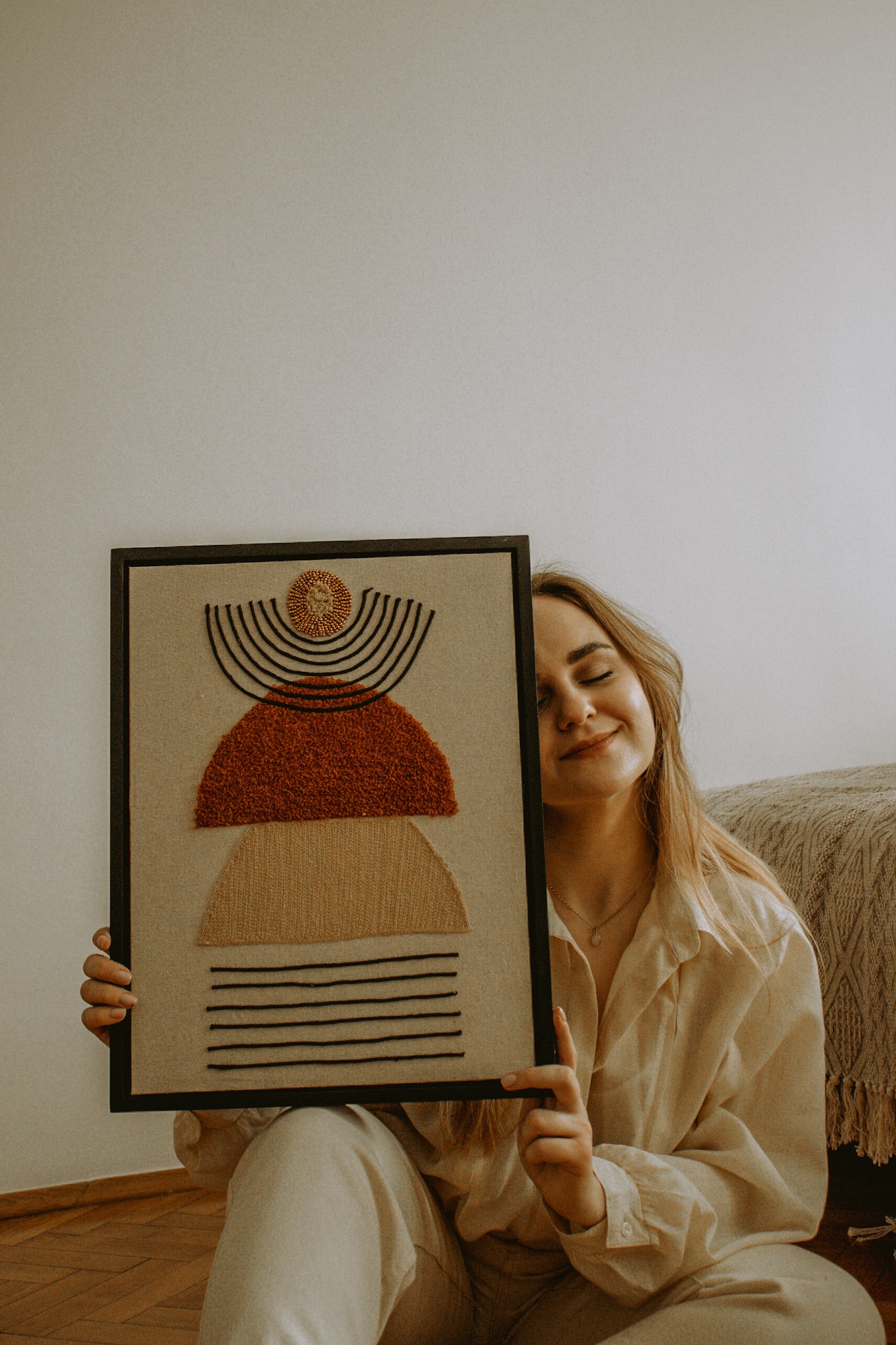 Kobieta z zamkniętymi oczami trzyma ręcznie robioną ramkę z abstrakcyjną kompozycją z tkanin i sznurków w odcieniach beżu, brązu i czerni, na tle beżowej ściany i fragmentu sofy.