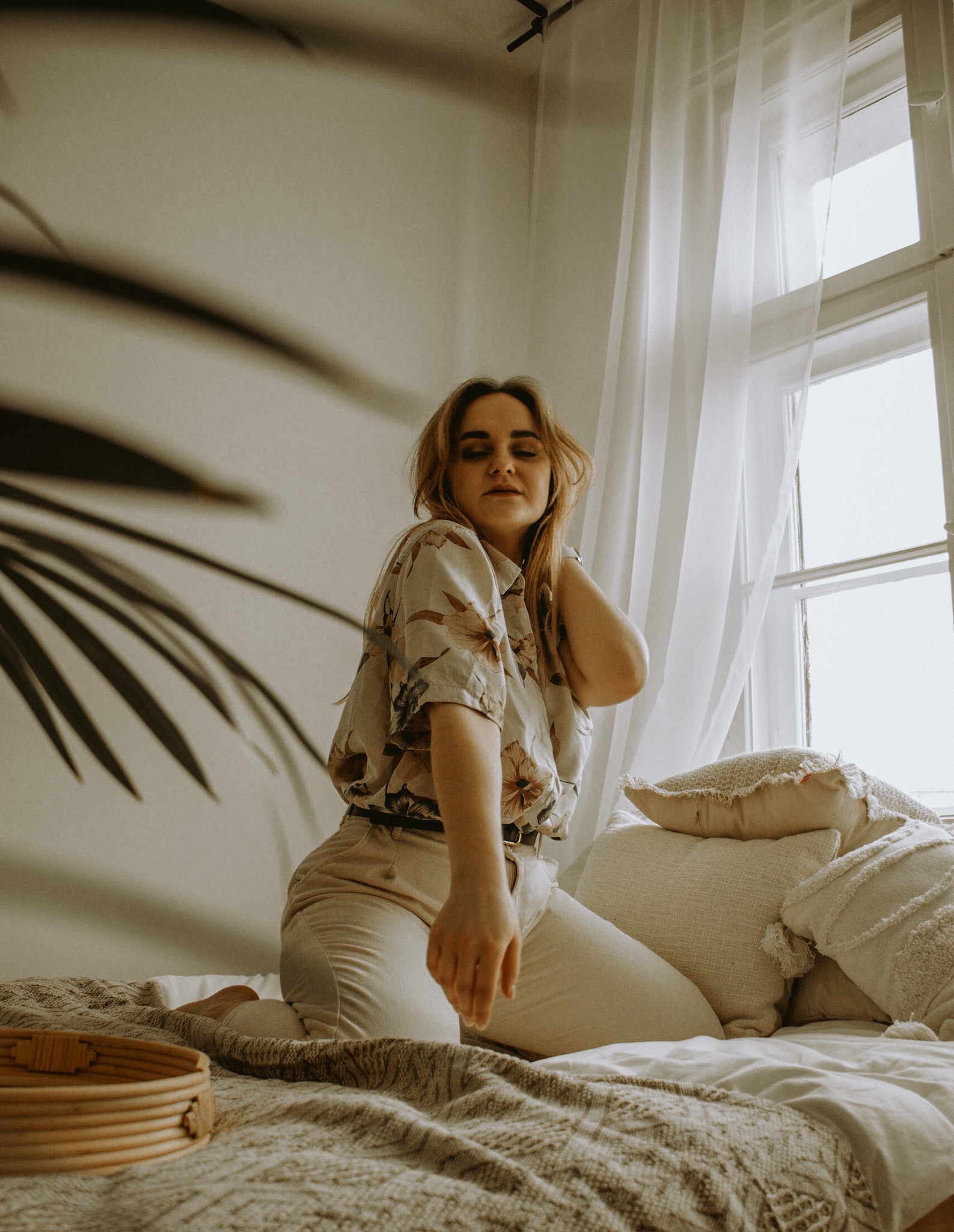 Kobieta w kwiecistej koszuli i beżowych spodniach pozuje na łóżku z poduszkami w naturalnym świetle w pobliżu okna z delikatnymi zasłonami, z widocznym fragmentem rośliny w tle.