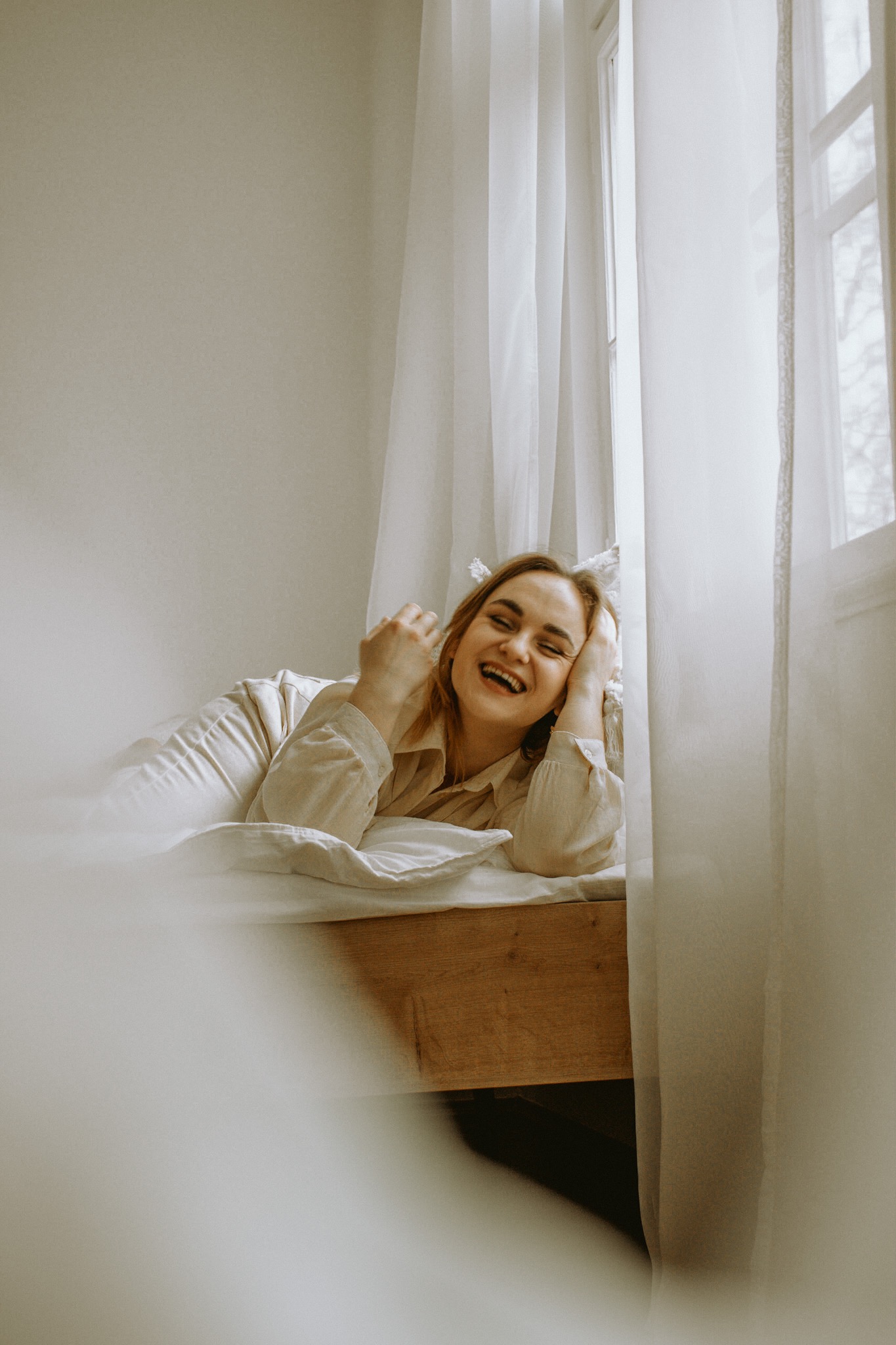 Uśmiechnięta kobieta w beżowej koszuli leży na łóżku obok okna z białymi zasłonami, naturalne światło.