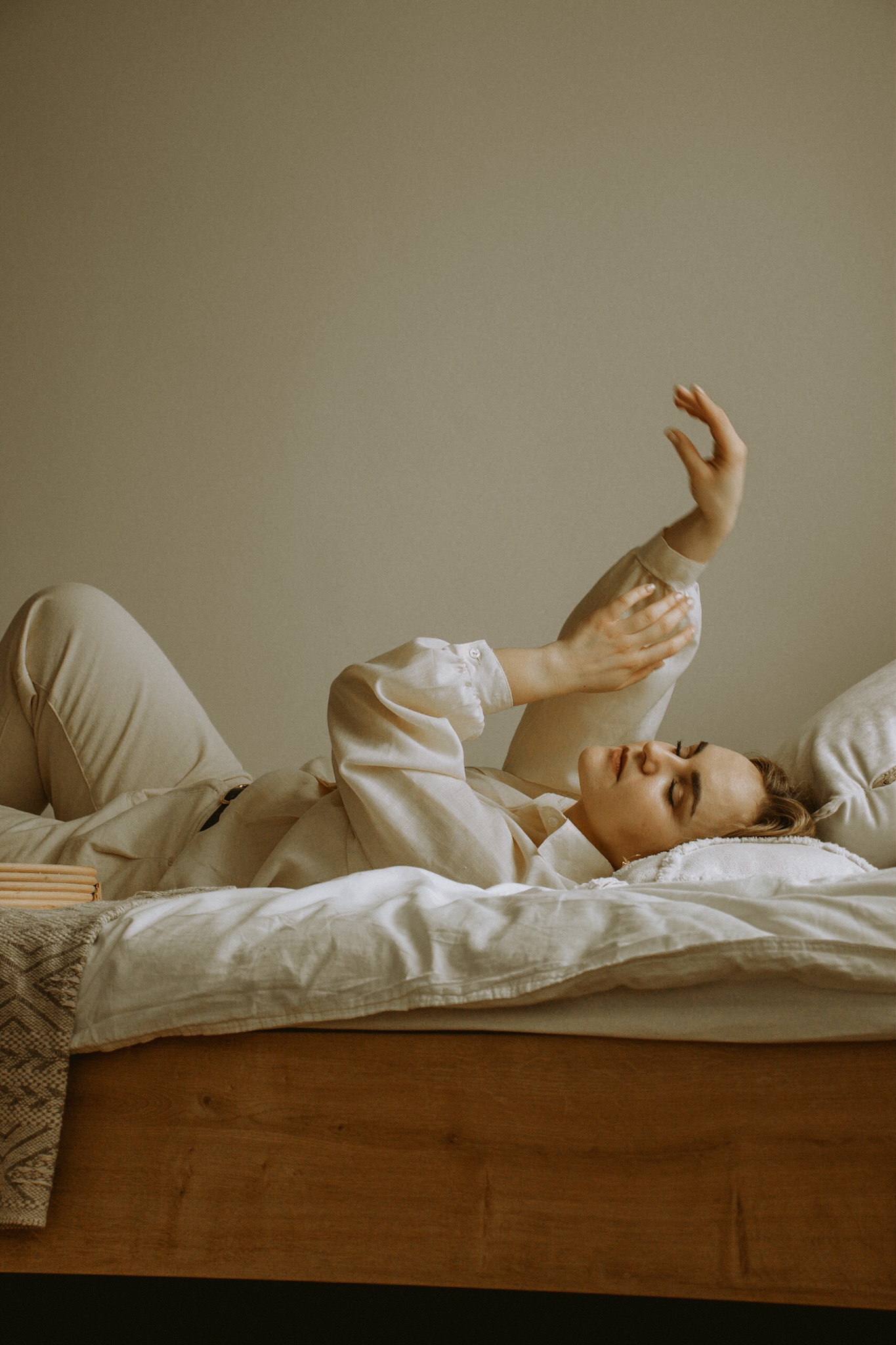 Kobieta w beżowym ubraniu leży na łóżku z podniesioną ręką, delikatne światło, minimalistyczne wnętrze.
