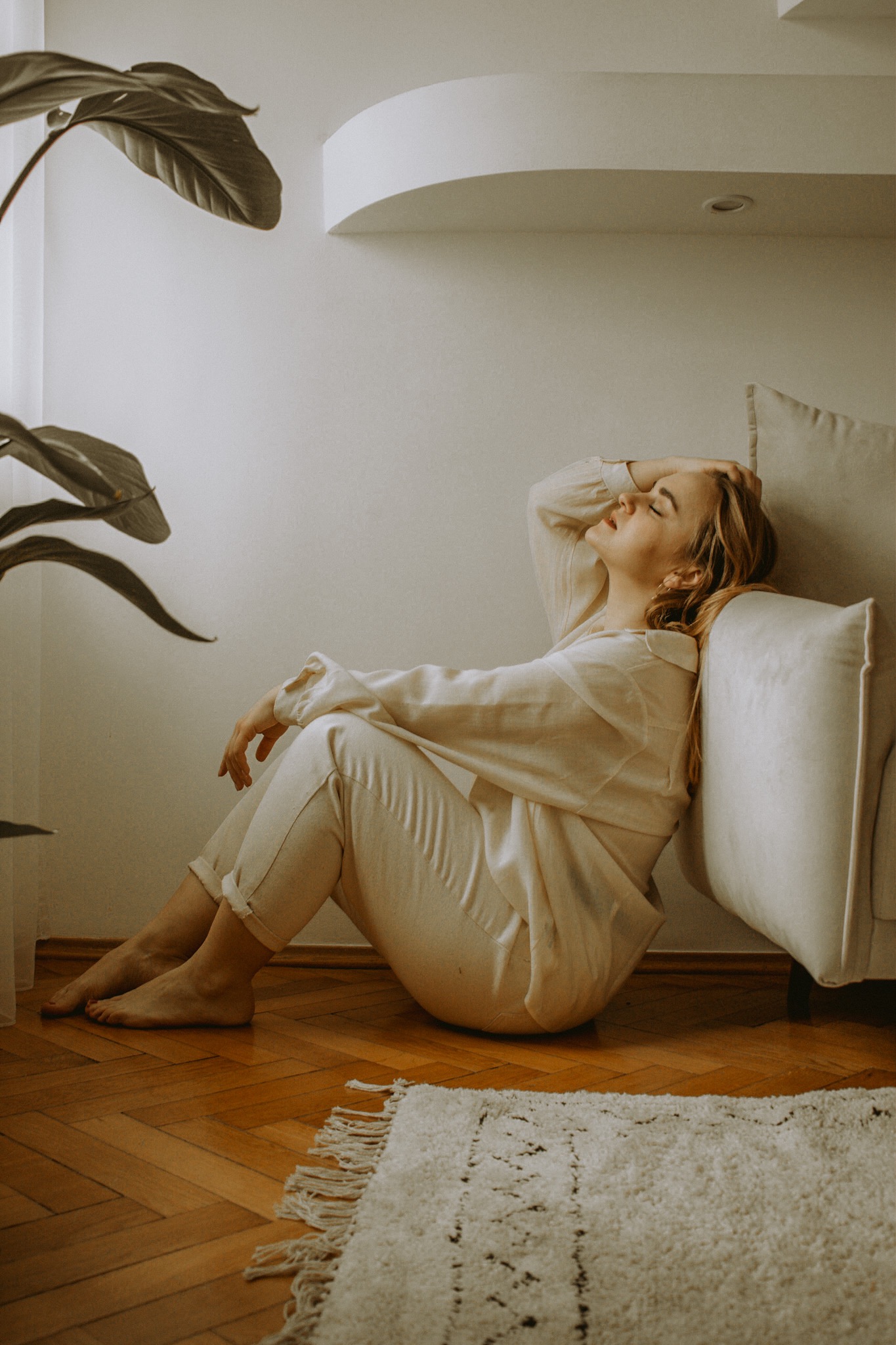 Kobieta w jasnym stroju siedzi oparta o poduszkę na drewnianej podłodze, z zamkniętymi oczami i uniesioną ręką, obok rośliny doniczkowej i dywanu z frędzlami.