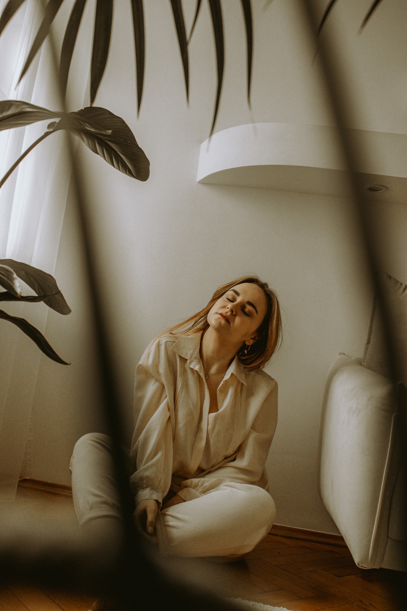 Kobieta siedząca na podłodze w jasnym wnętrzu, z zamkniętymi oczami i głową odchyloną do tyłu, w tle roślina doniczkowa i minimalistyczna półka.