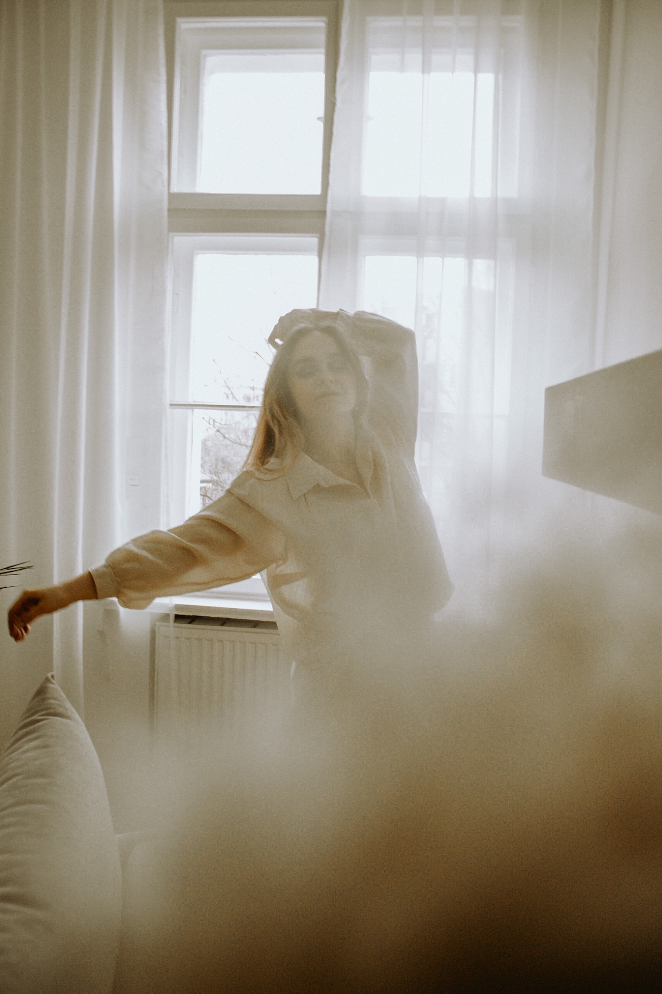 Kobieta w beżowej koszuli pozuje przy oknie z białymi zasłonami, rozmyta sylwetka z przodu kadru.