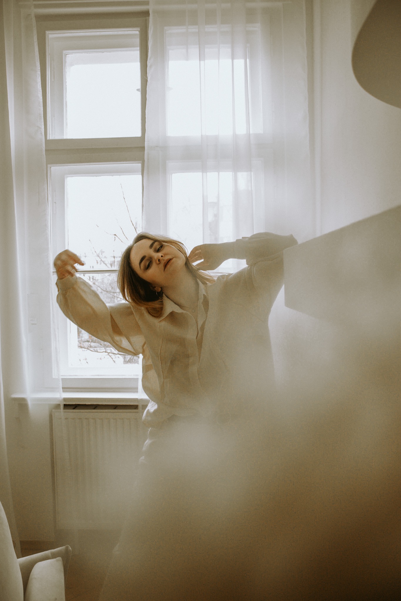 Kobieta w jasnej koszuli pozuje przy oknie z białymi firankami, delikatne światło, artystyczne ujęcie z rozmytym pierwszym planem.