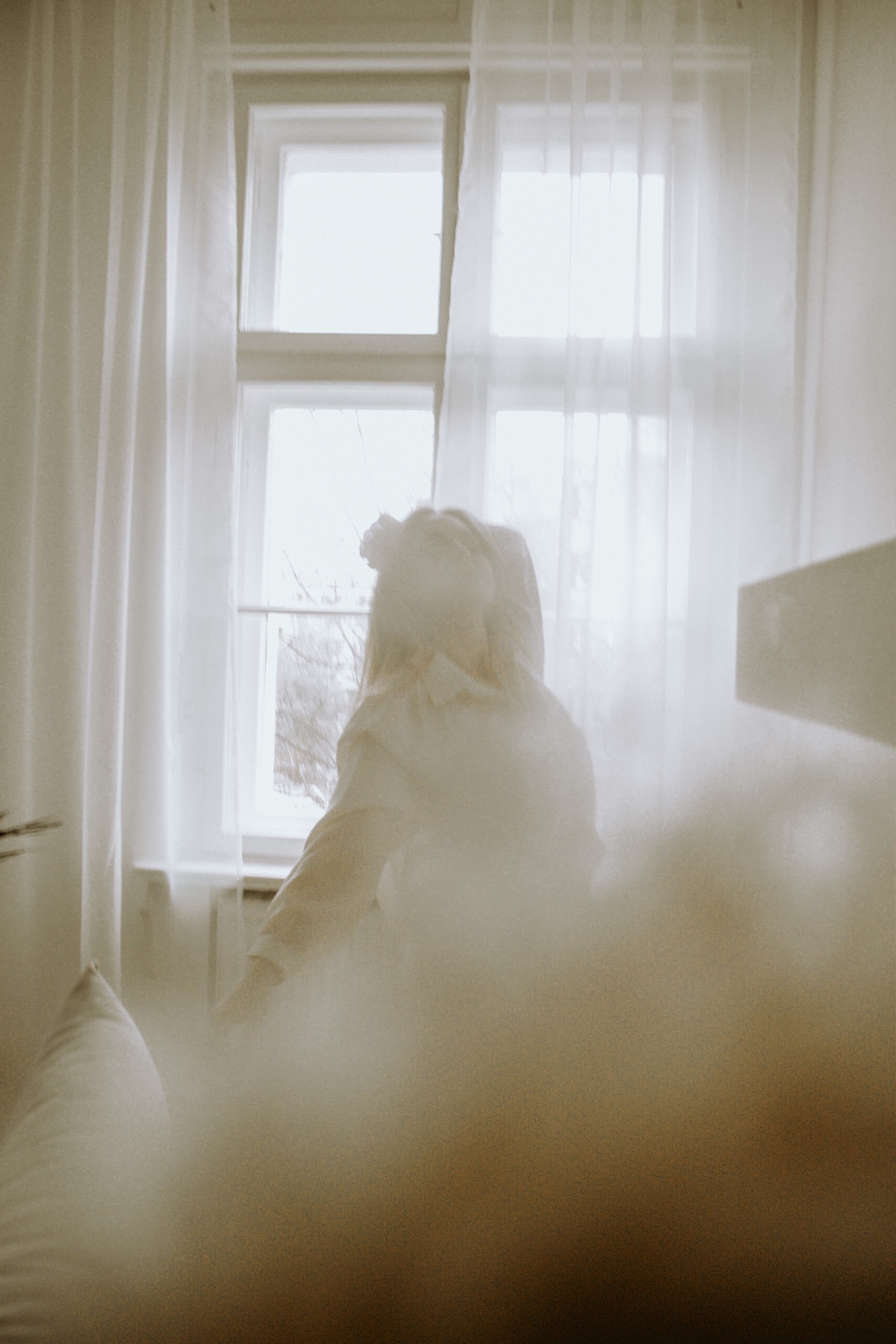 Kobieta w jasnej koszuli stoi w pokoju, częściowo zasłonięta miękkim, rozmytym efektem, z oknem w tle przysłoniętym białymi firankami.